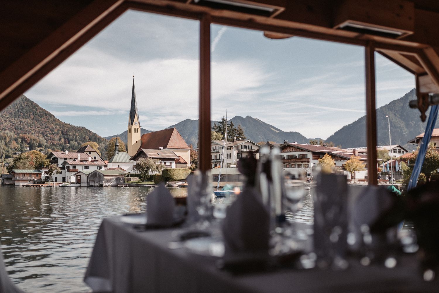 View of Hallstatt village and church spire across the lake through a restaurant window with table settings in foreground.