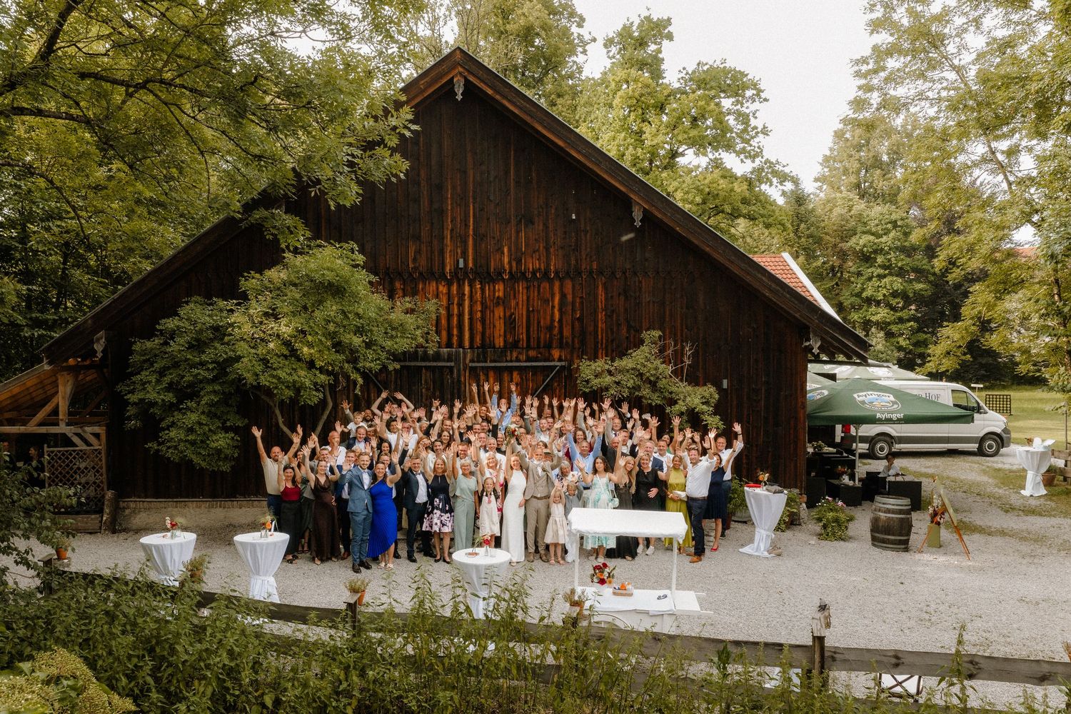 Large group gathered outside a rustic barn venue surrounded by green trees.