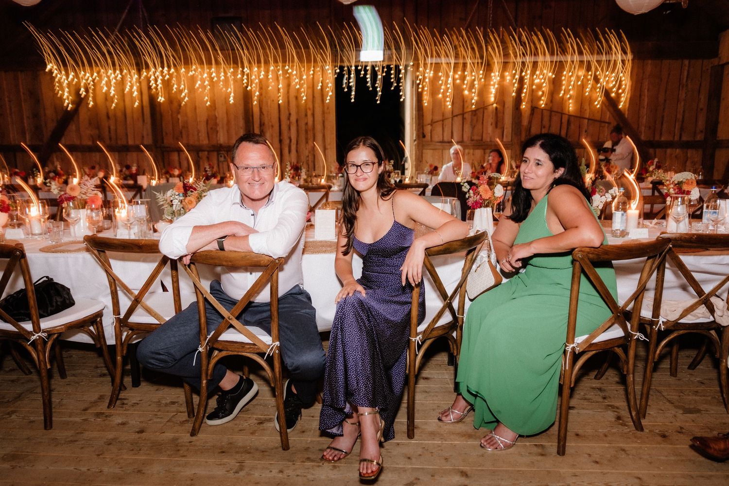 Wedding guests seated at long tables under warm string lights in rustic barn setting.