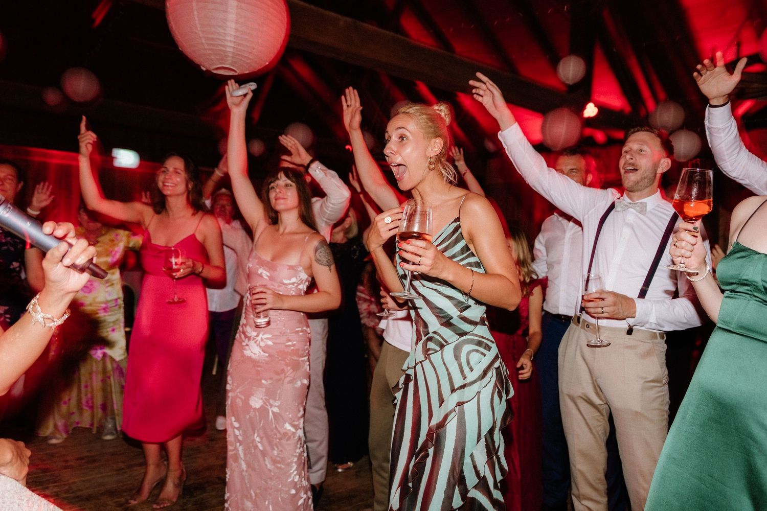 Wedding guests dance energetically under red lighting and paper lanterns at a lively reception celebration.
