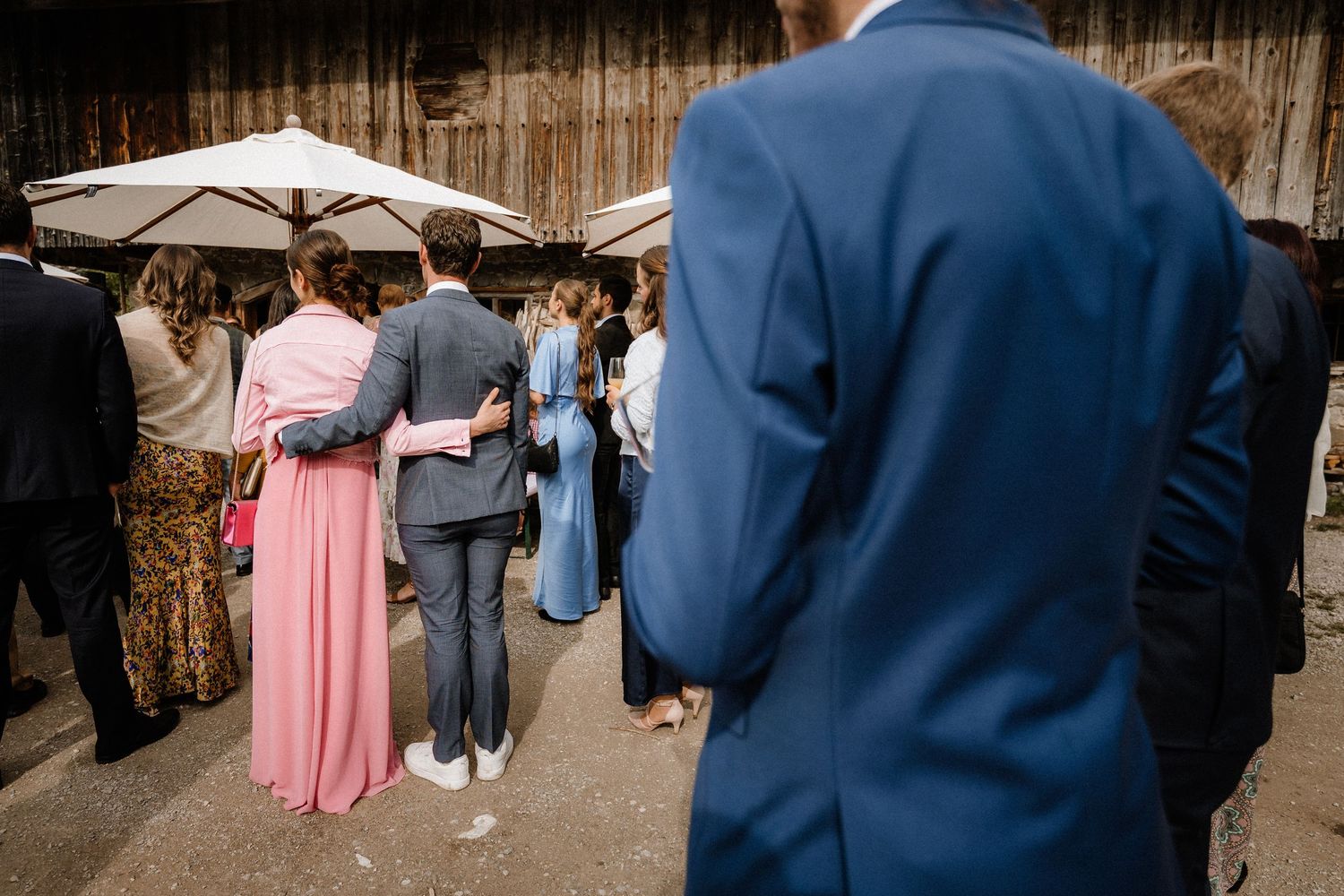 Wedding guests in formal attire gathered under white umbrellas at an outdoor venue.