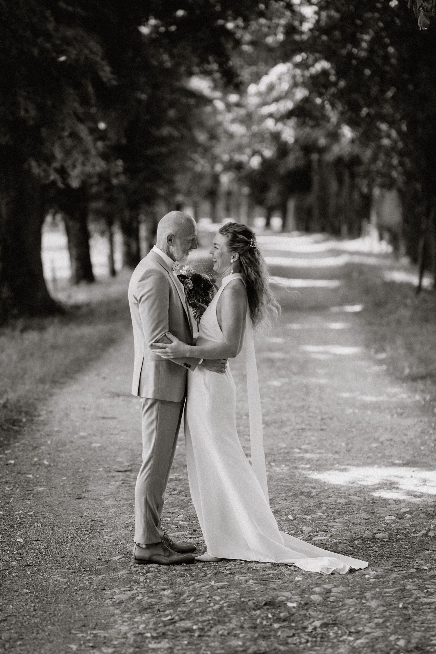 A black and white photo captures a romantic wedding moment on a tree-lined path.
