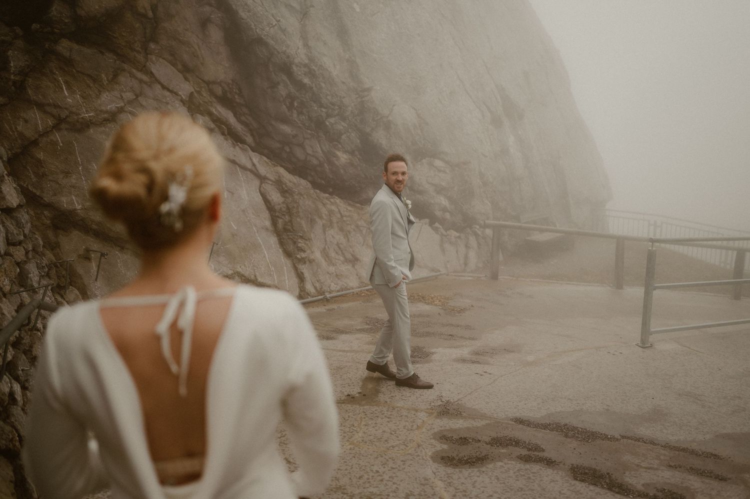Couple in white wedding attire walking along a misty mountain path.