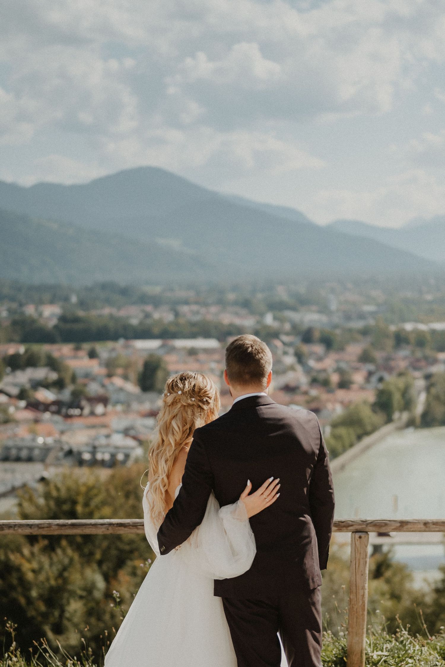 Newlyweds embrace while overlooking a scenic mountain valley town from a viewing platform.