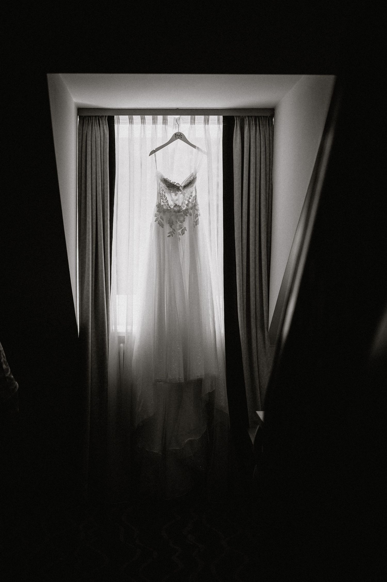 Ein weißes Hochzeitskleid hängt elegant vor einem Fenster mit dunklen Vorhängen, die eine dramatische Silhouette schaffen. Dama Fotografie