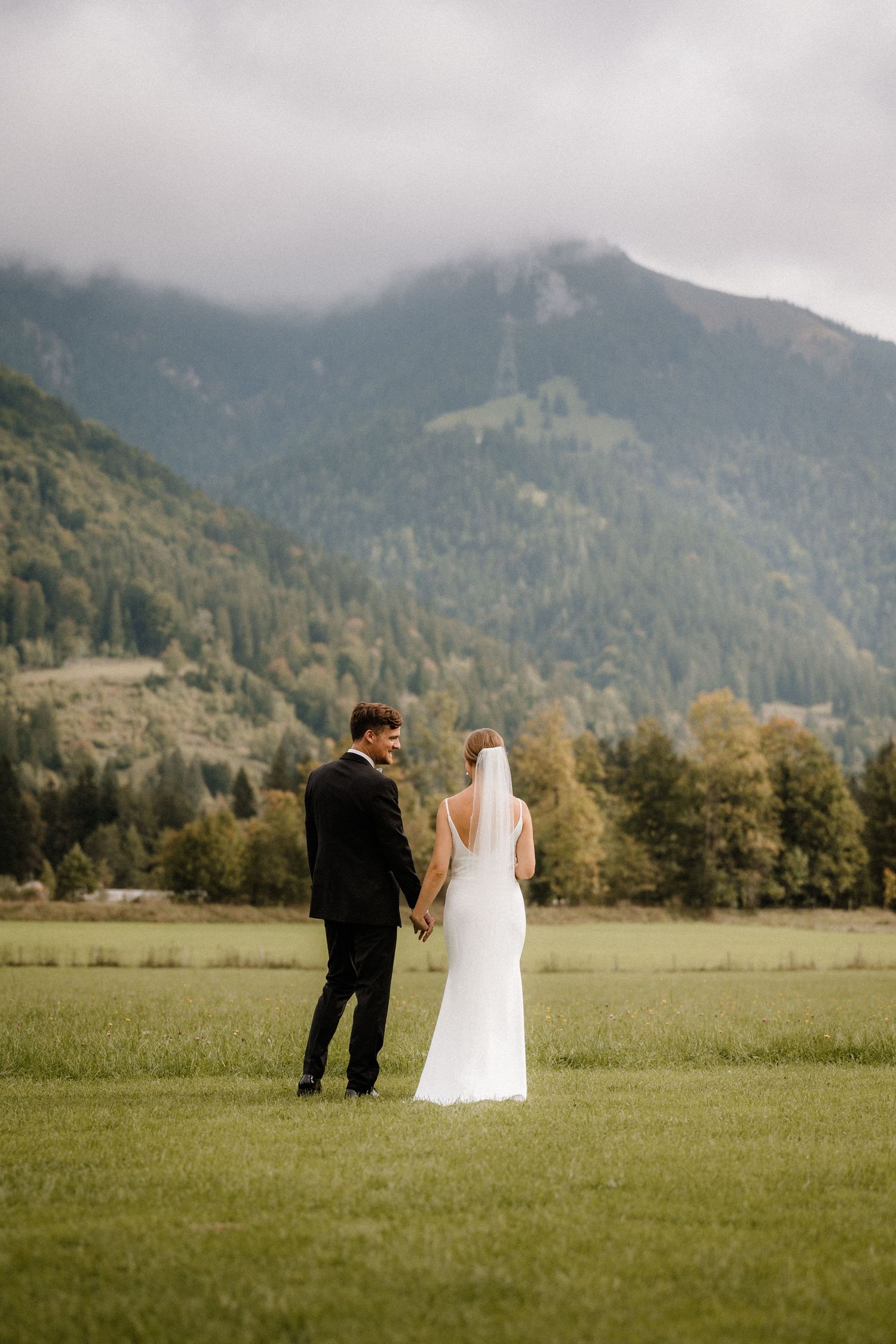 A bride and groom walk hand in hand across a meadow with mountains in the background.