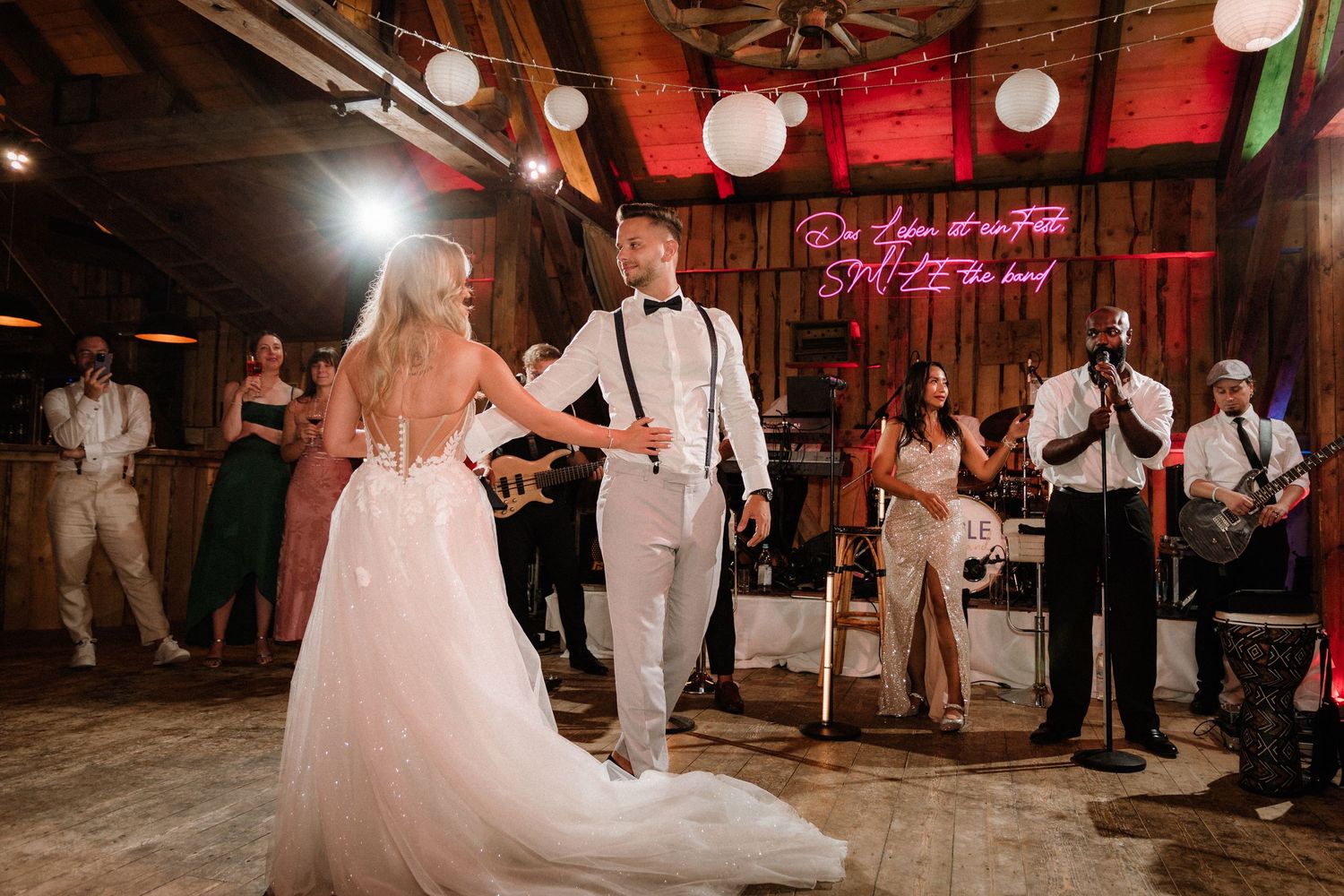Wedding party dancing in a rustic barn venue illuminated by warm lighting and hanging lanterns.