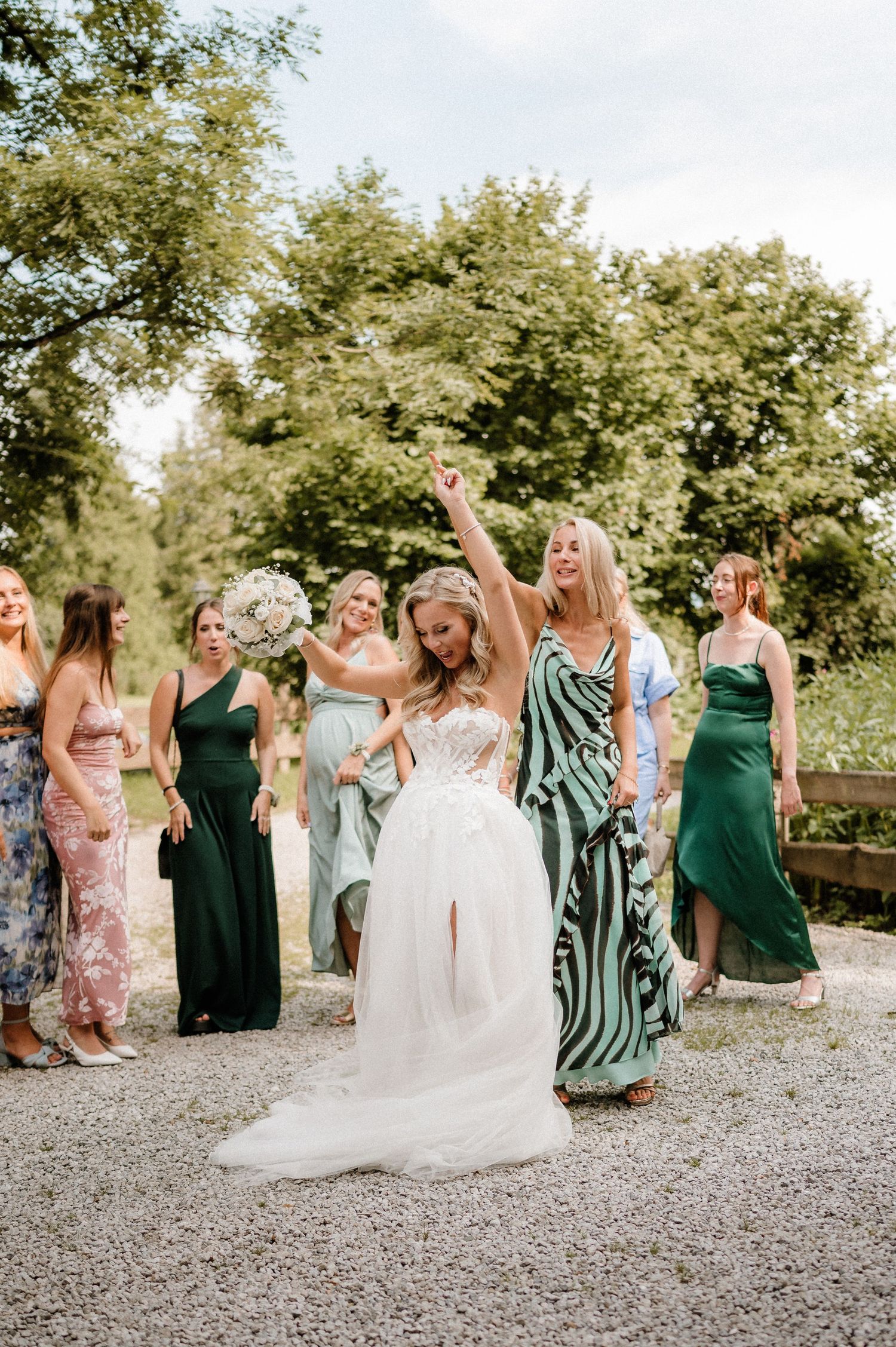Wedding party in green dresses and floral prints dancing and celebrating on a gravel path.
