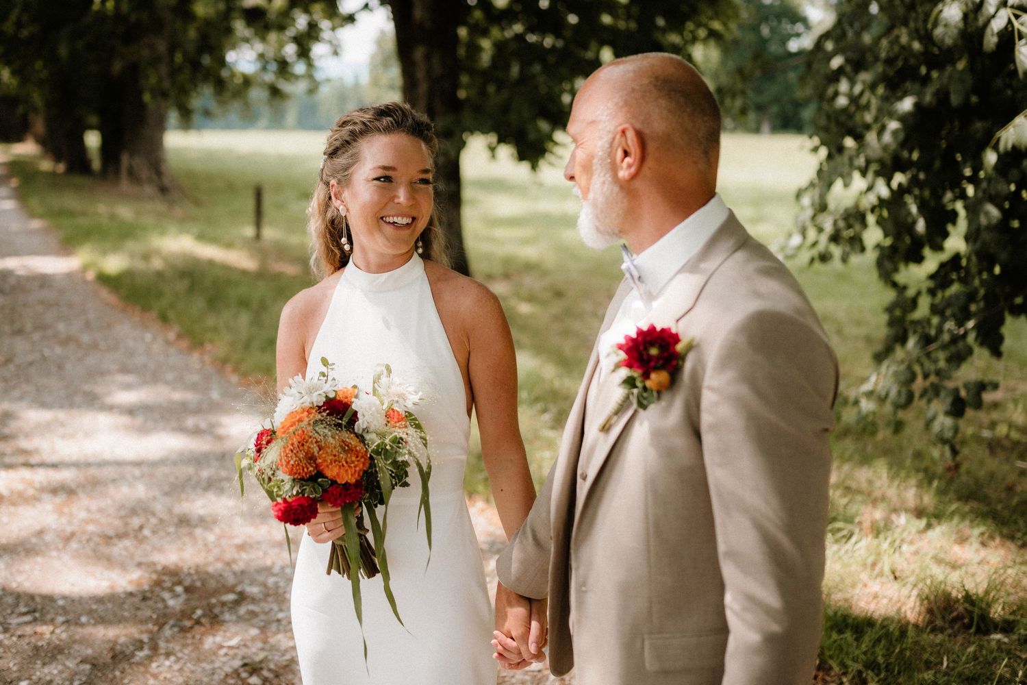 Newlyweds share joyful moments on a sunlit path with colorful bouquet and tan suit.