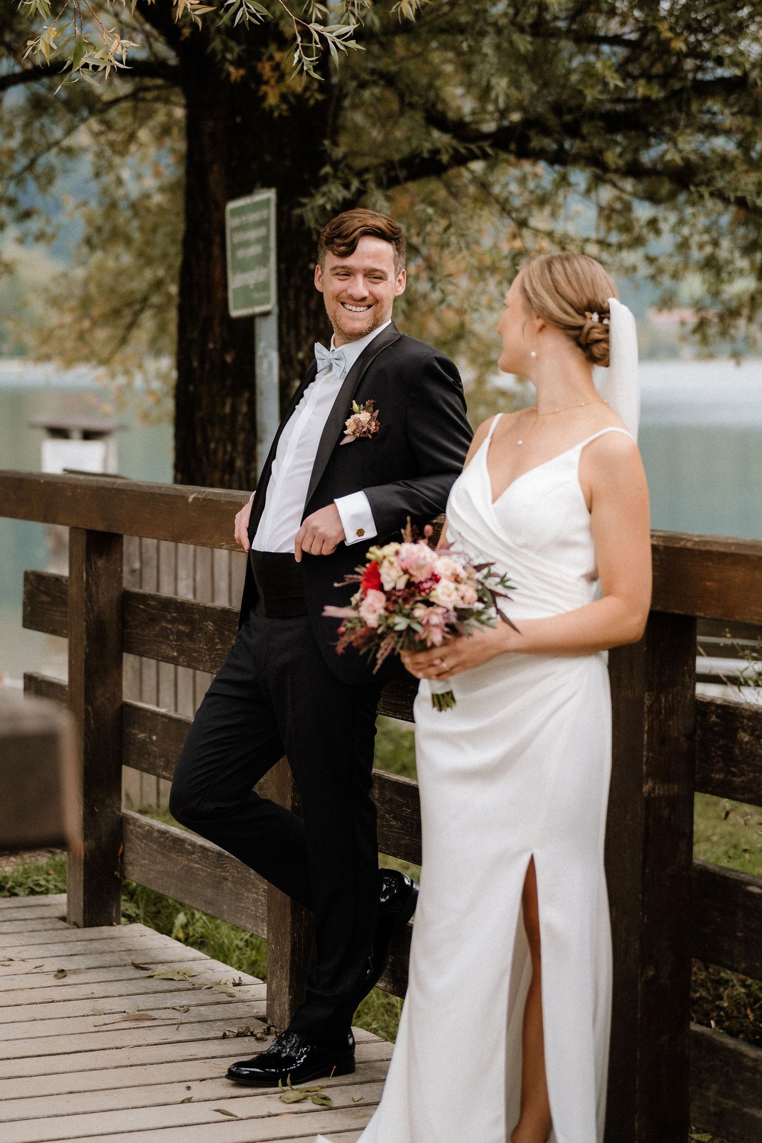 Wedding couple poses on wooden deck by lake with autumn trees.