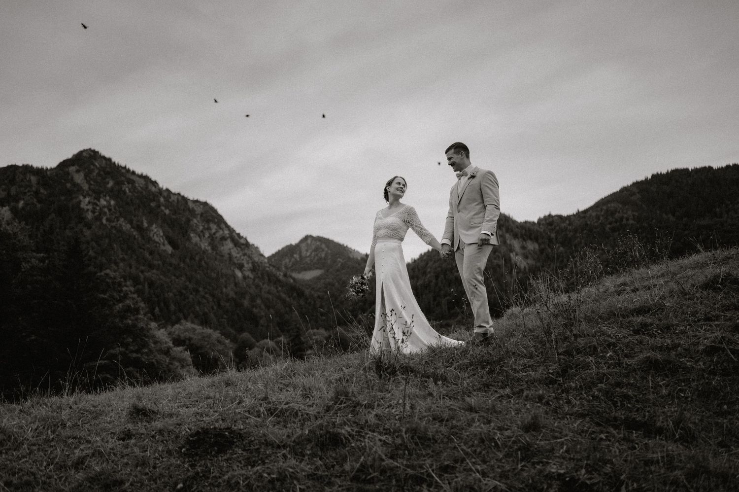 Das Hochzeitspaar erkundet eine dramatische Berglandschaft unter bewölktem Himmel. Von Dama Fotografie Bad Tölz