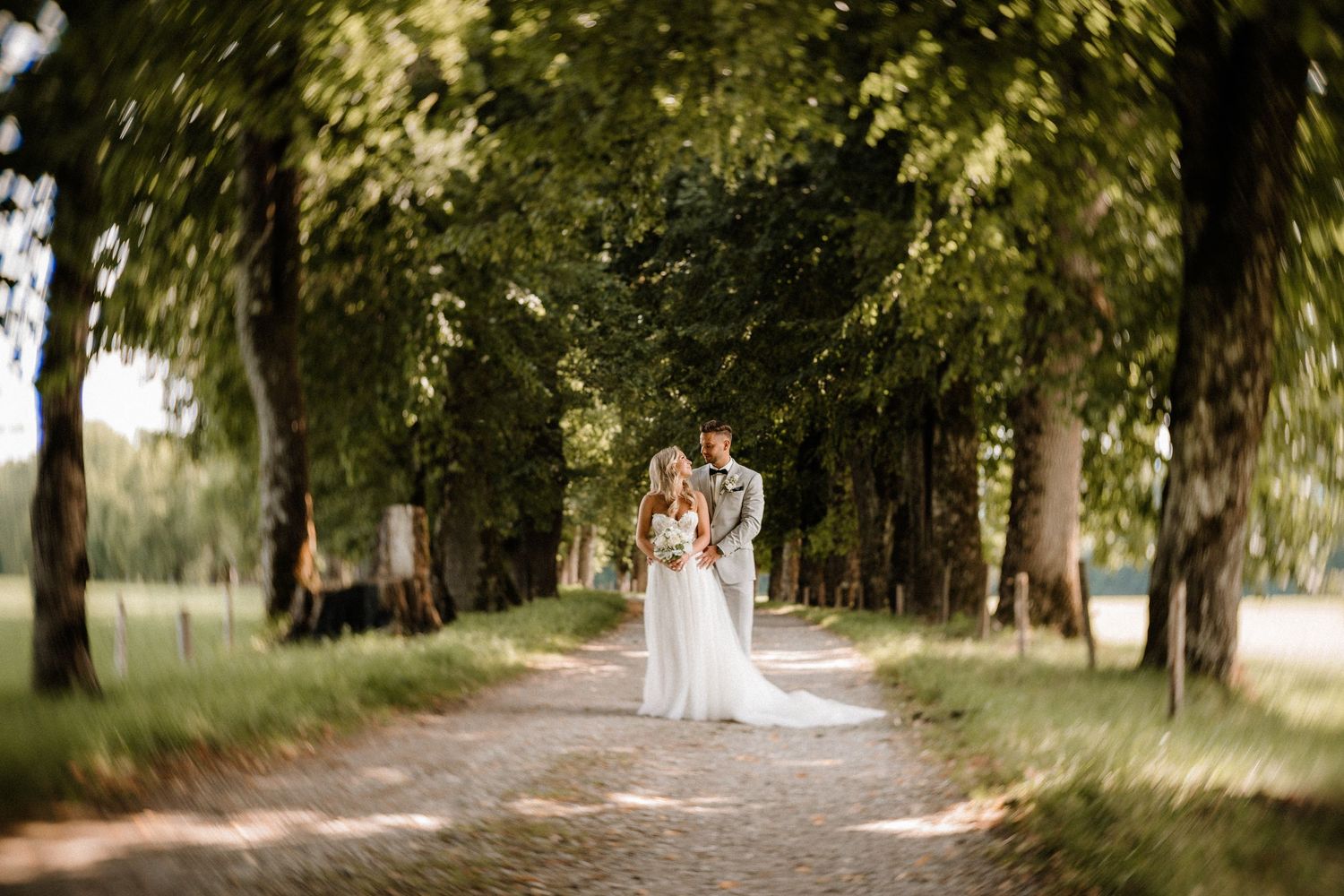 A couple in wedding attire stands together on a tree-lined path in a romantic outdoor setting.