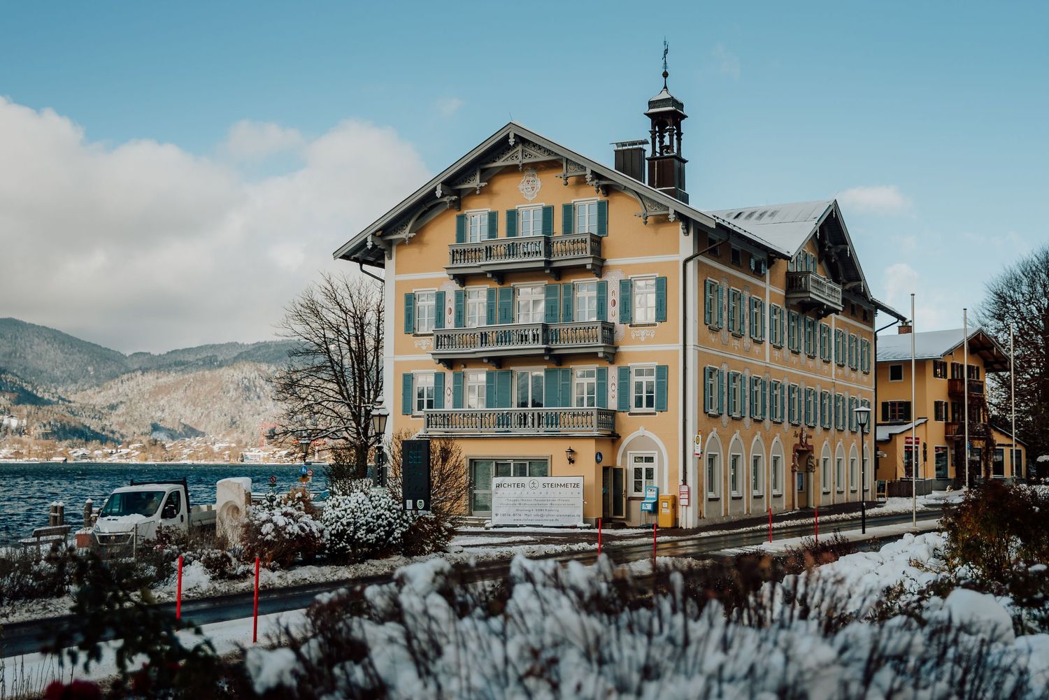Traditional Alpine hotel building with yellow facade stands in snowy winter landscape overlooking mountain lake in Bavaria.