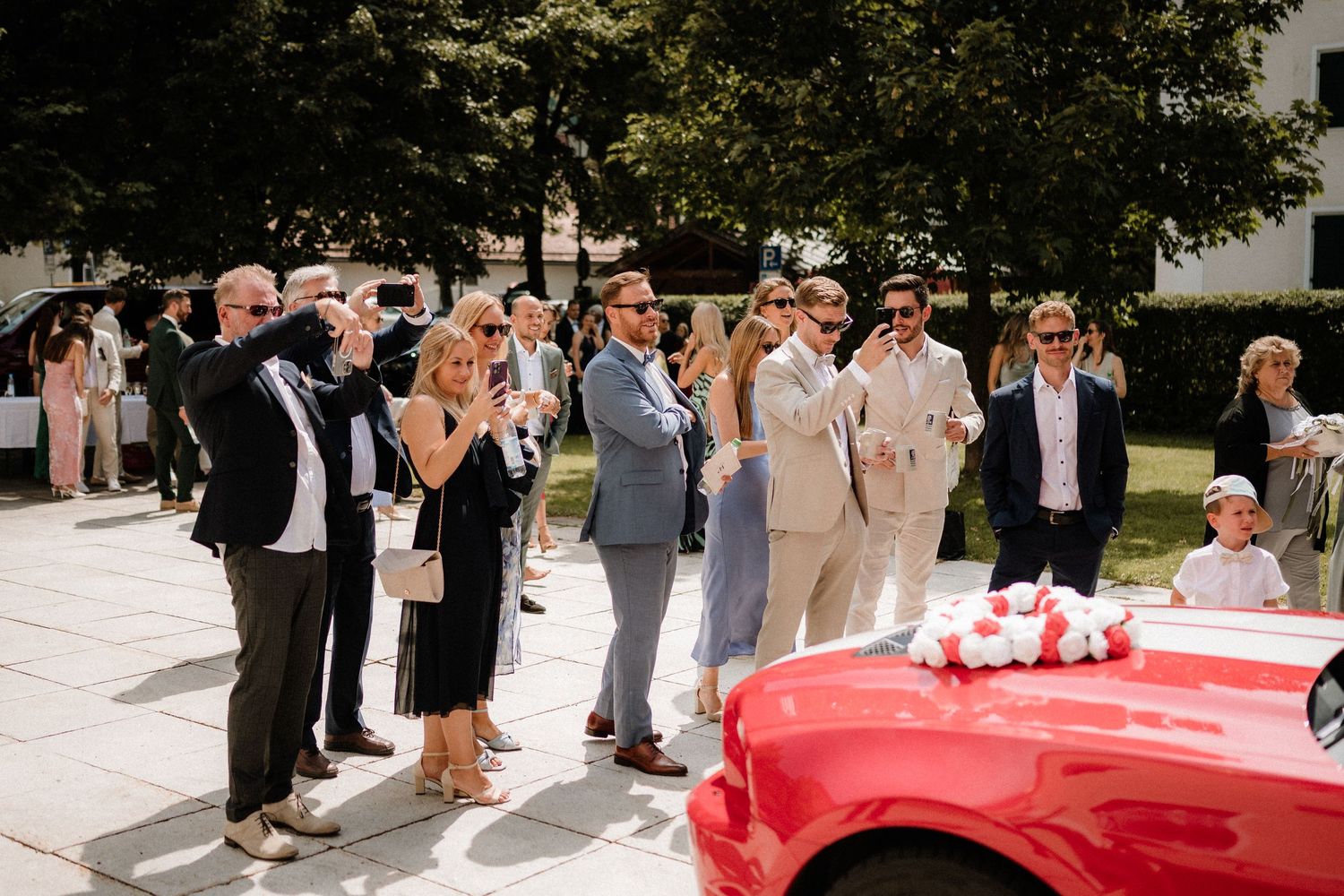 Wedding guests gathering around a bright red vintage car parked outdoors.