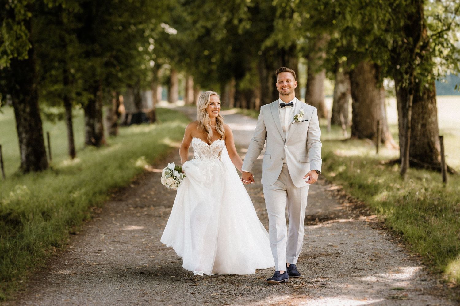 Das Hochzeitspaar geht in einem eleganten weißen Kleid und einem hellen Anzug auf einem von Bäumen geseilten Weg entlang. Von Dama Fotograf