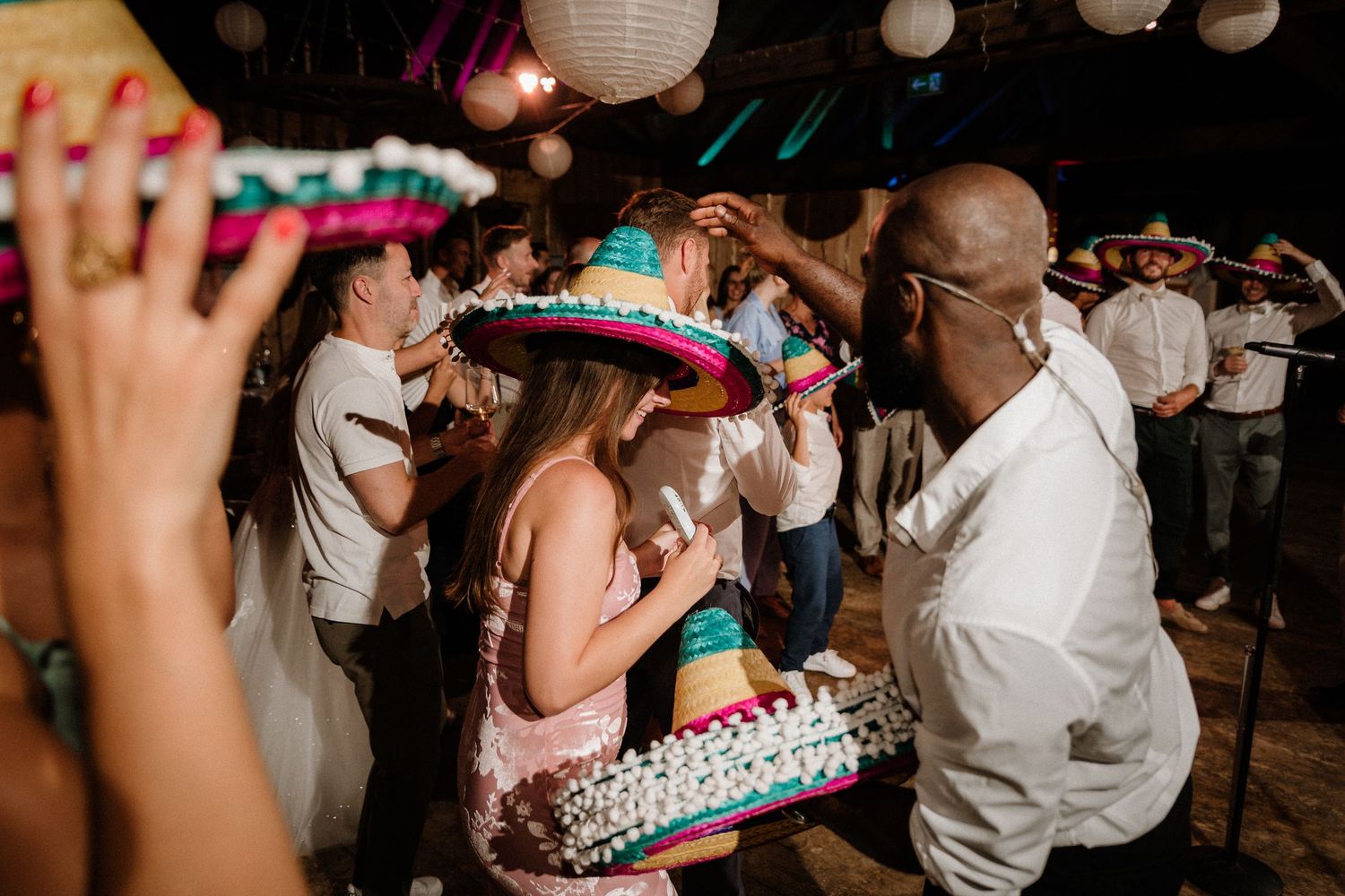 Wedding guests dancing while wearing colorful Mexican sombreros at an indoor celebration.