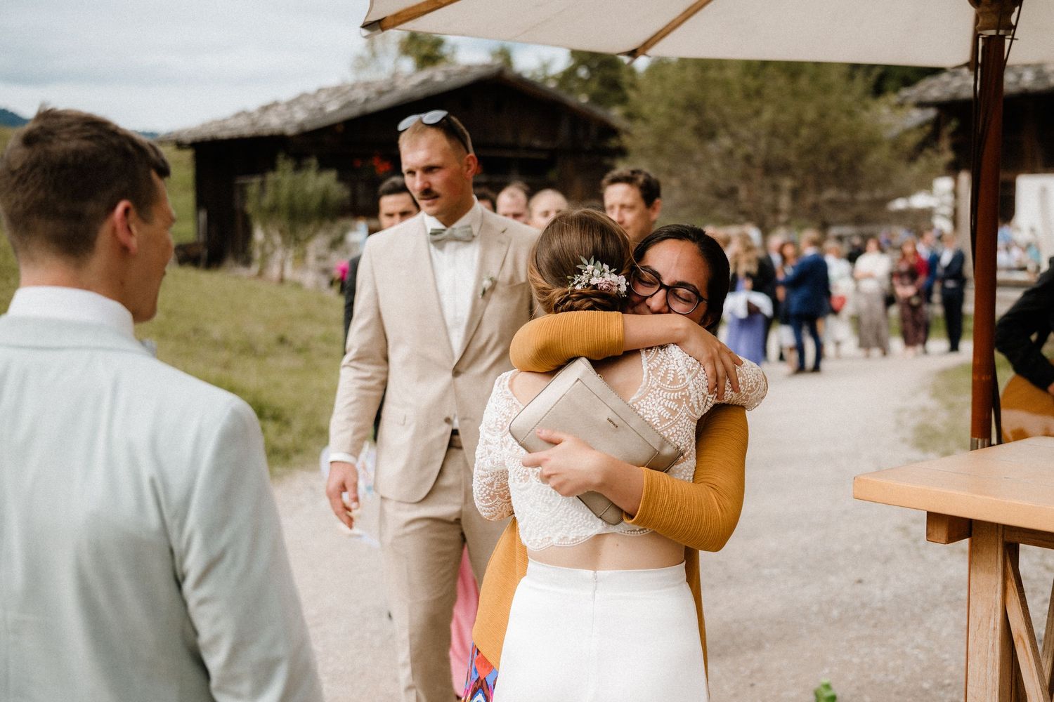Wedding guests socialize under white umbrellas on a gravel path near mountain scenery.