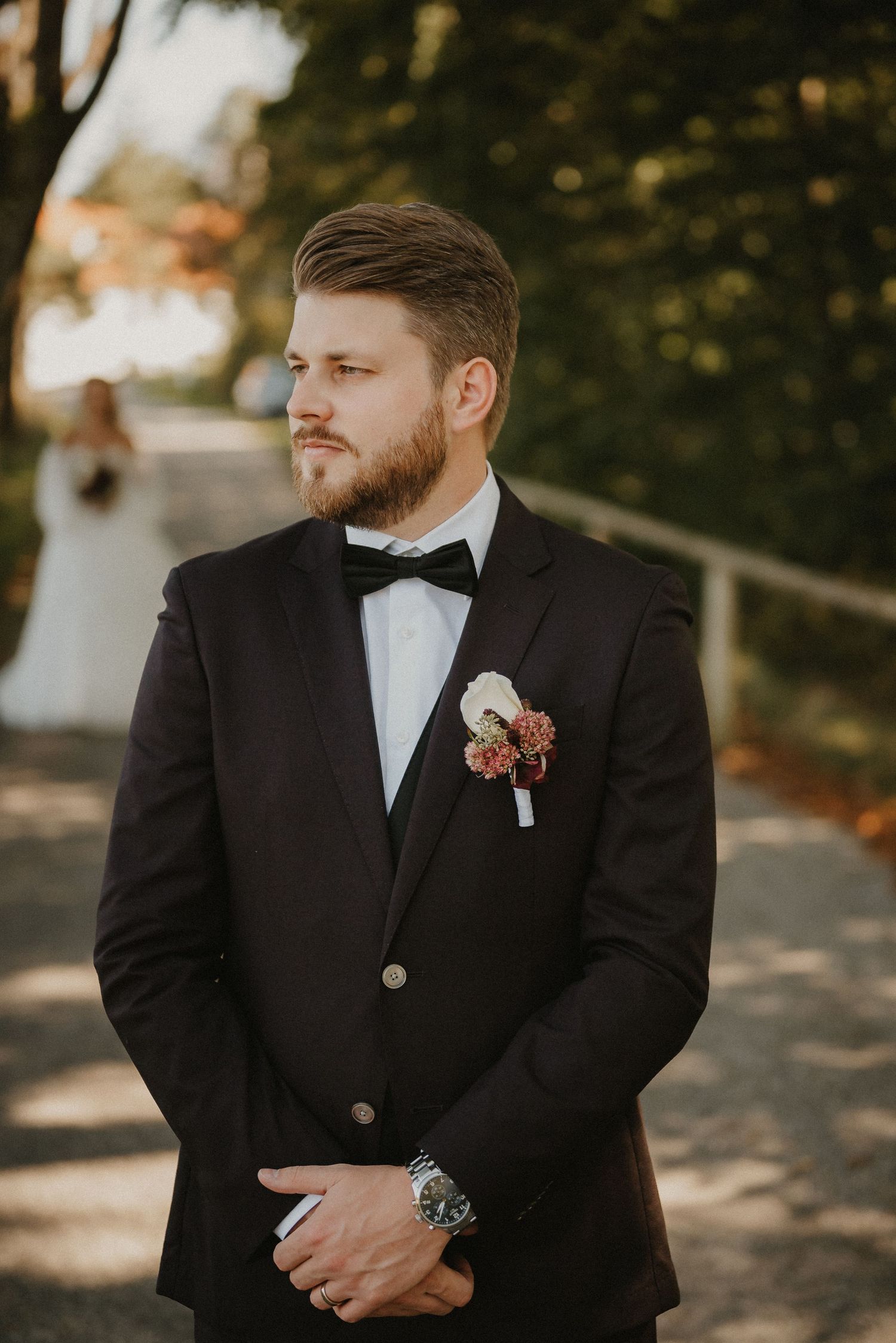 A groom in a classic black tuxedo and bow tie poses outdoors with a floral boutonniere.