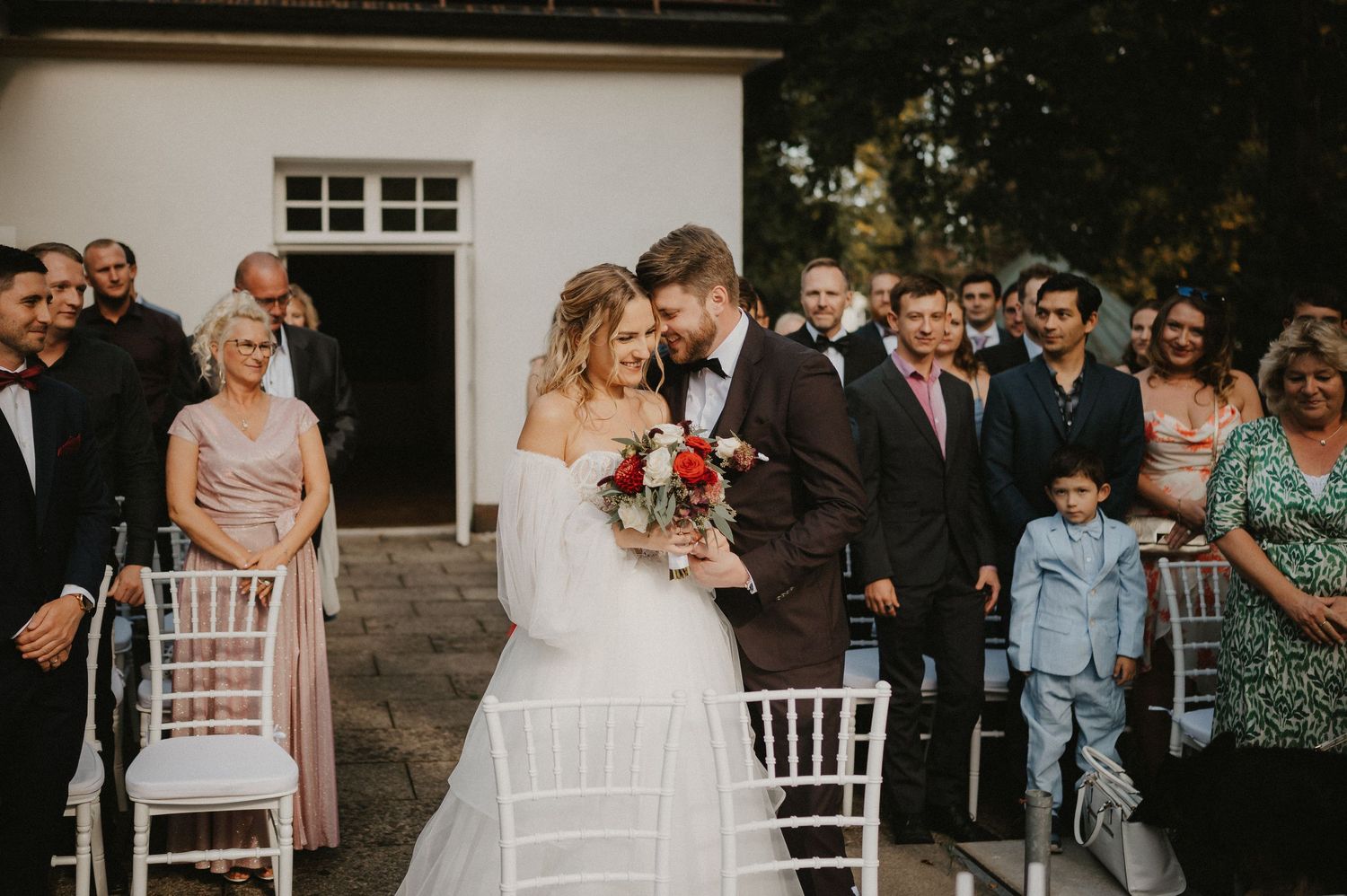 Wedding guests seated in rows of white chairs during an outdoor ceremony.