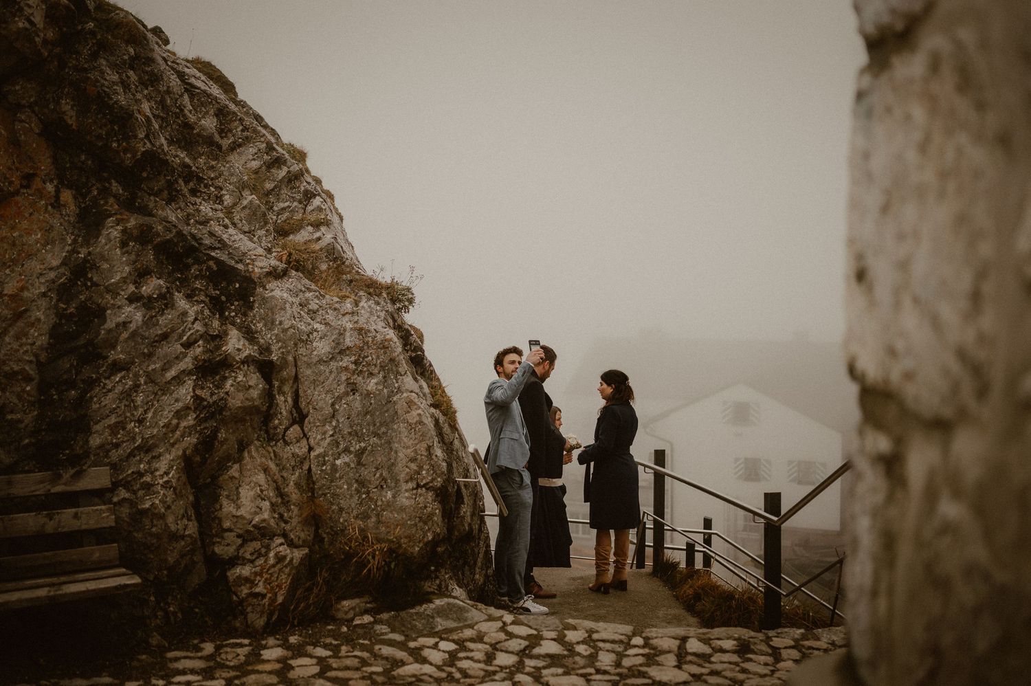 A couple shares an intimate moment on a misty mountain path with dramatic rocky cliffs in the background.