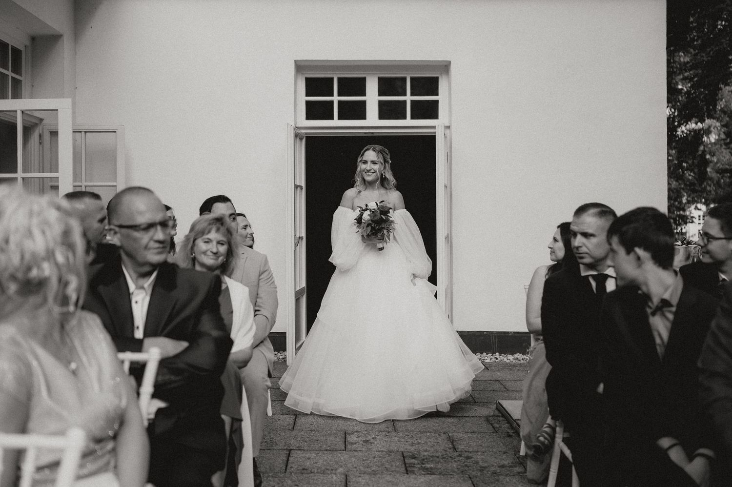 Wedding ceremony guests seated in white chairs watch the outdoor proceedings at a charming white building venue.