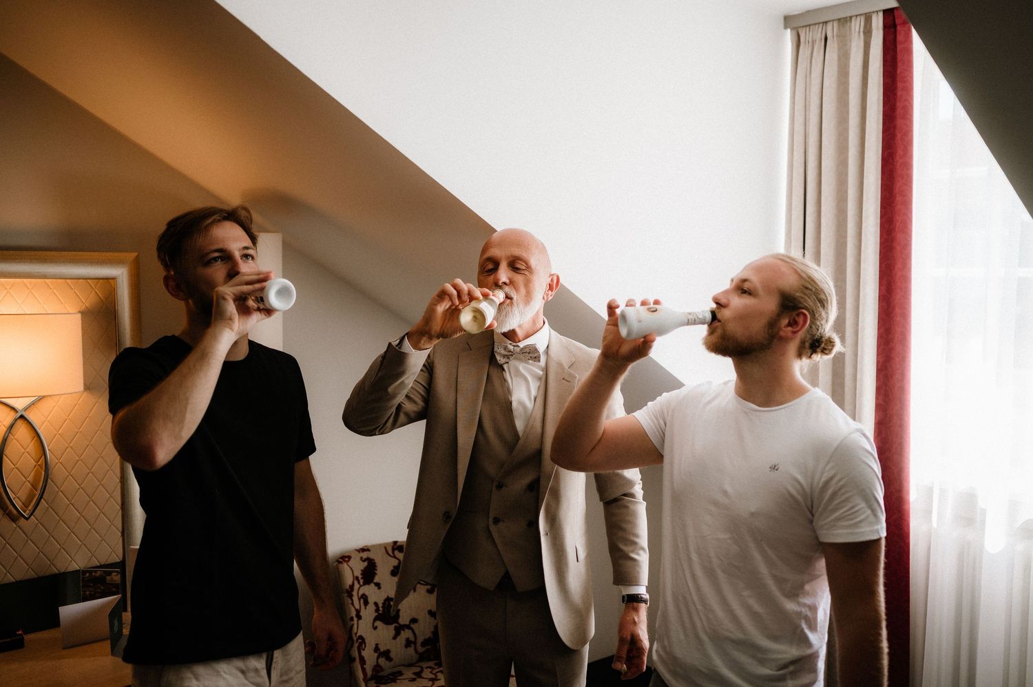 Wedding party members toast with drinks during pre-ceremony preparations in a sunlit room.