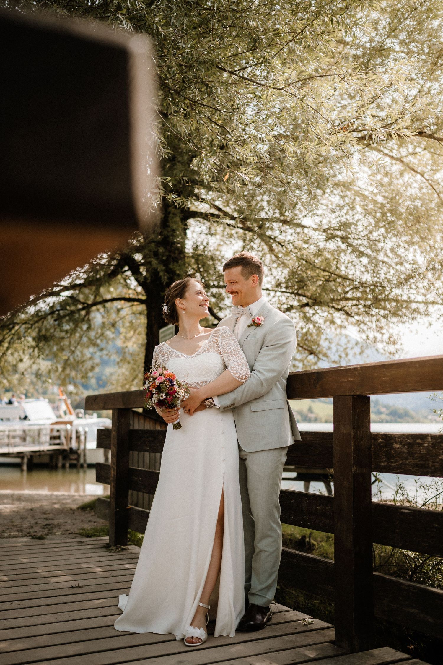 A joyful wedding couple shares a laugh on a wooden deck surrounded by trees.