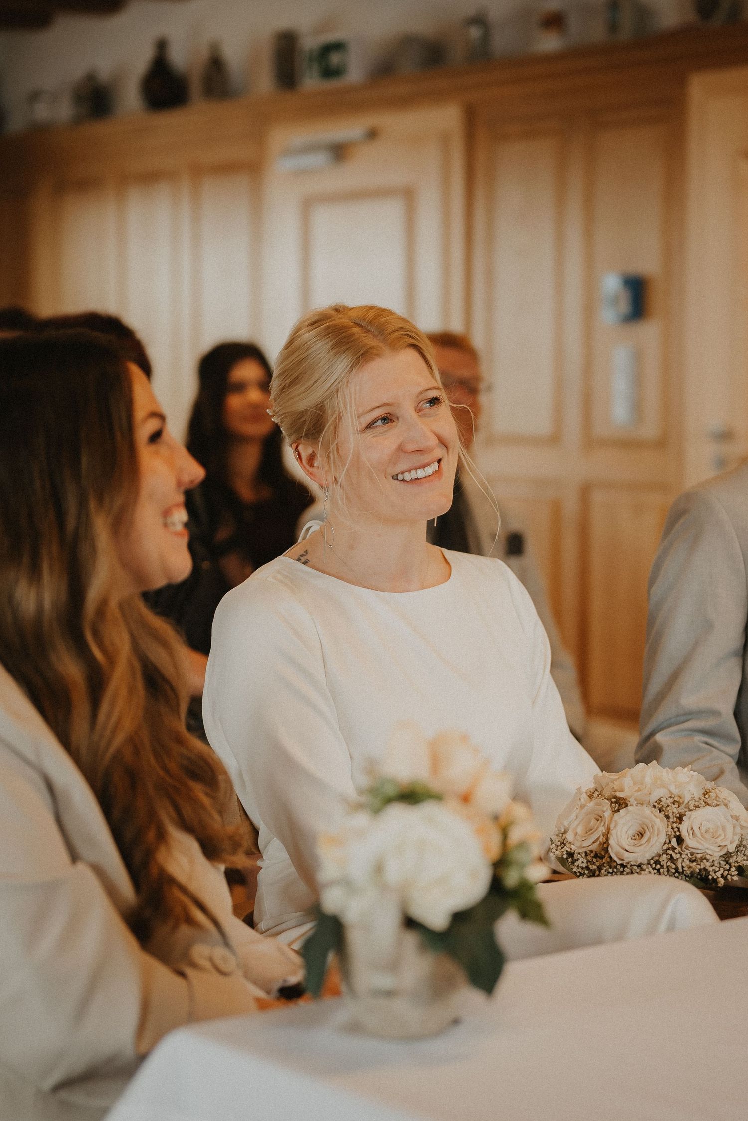 Wedding guests sharing joyful laughter during an intimate indoor celebration with white floral decorations.