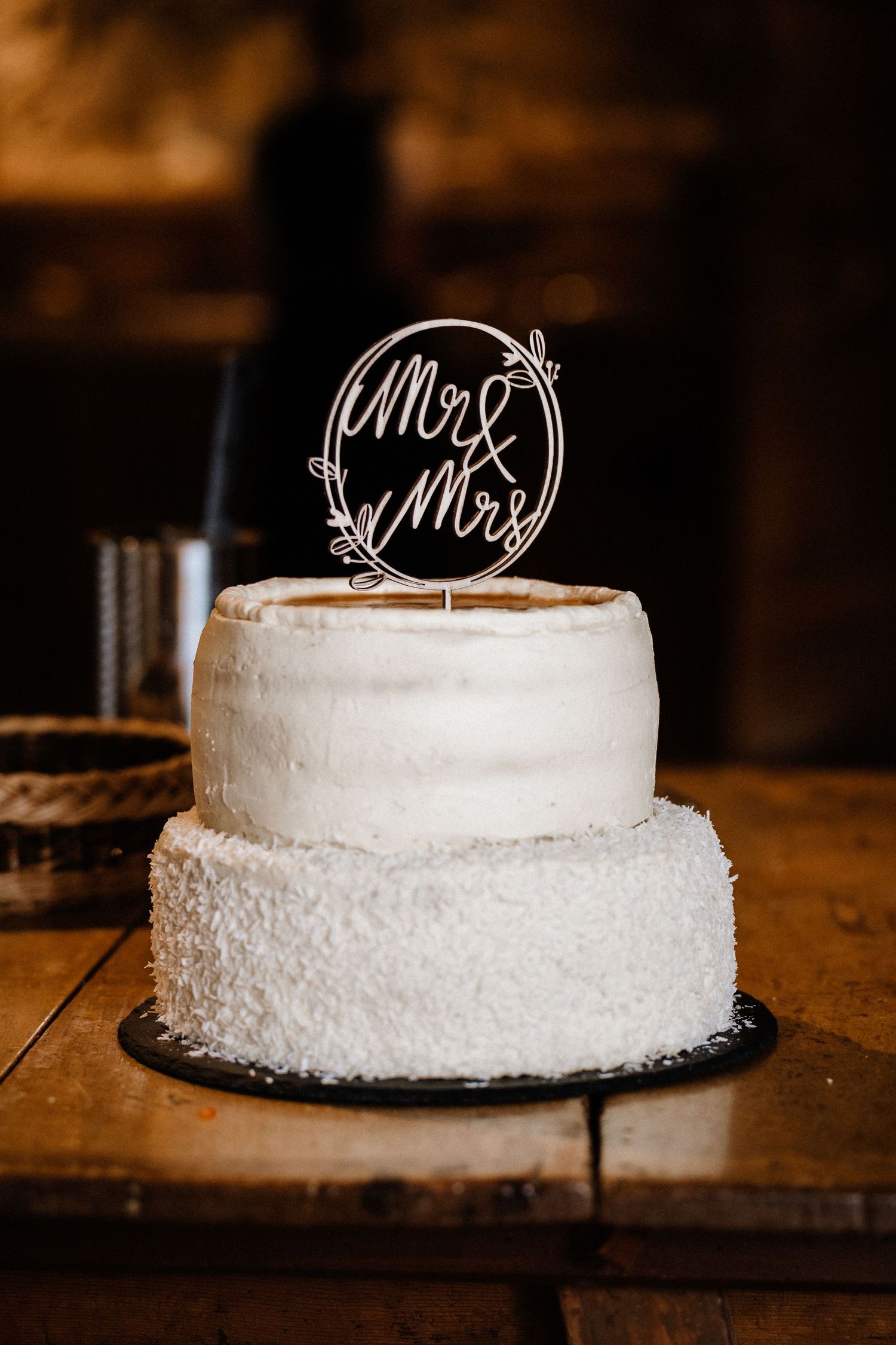 White tiered wedding cake with delicate frosting and elegant Mr and Mrs topper on rustic wooden table.