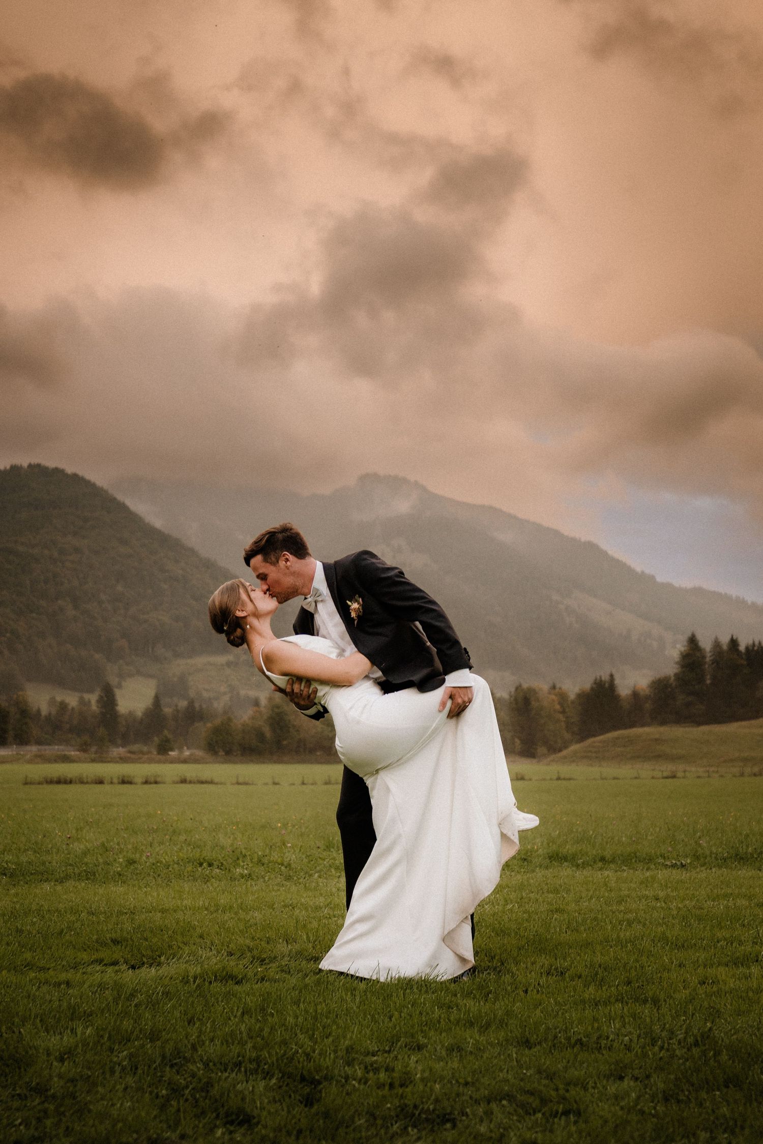 Couple in wedding attire dips dramatically in grassy field with mountains in background.