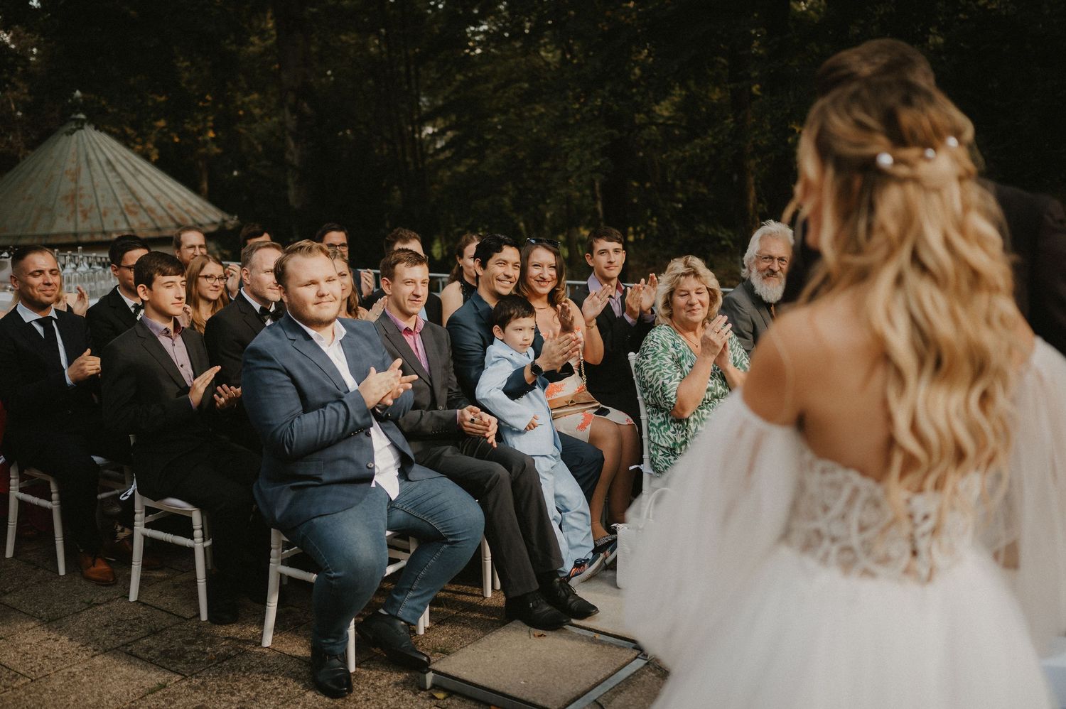 Wedding guests seated in white chairs during an outdoor ceremony surrounded by trees.