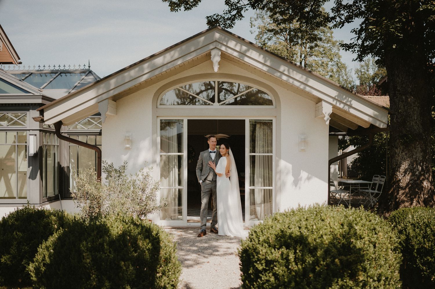 A couple stands in the doorway of a charming white garden cottage with arched entrance and manicured hedges.