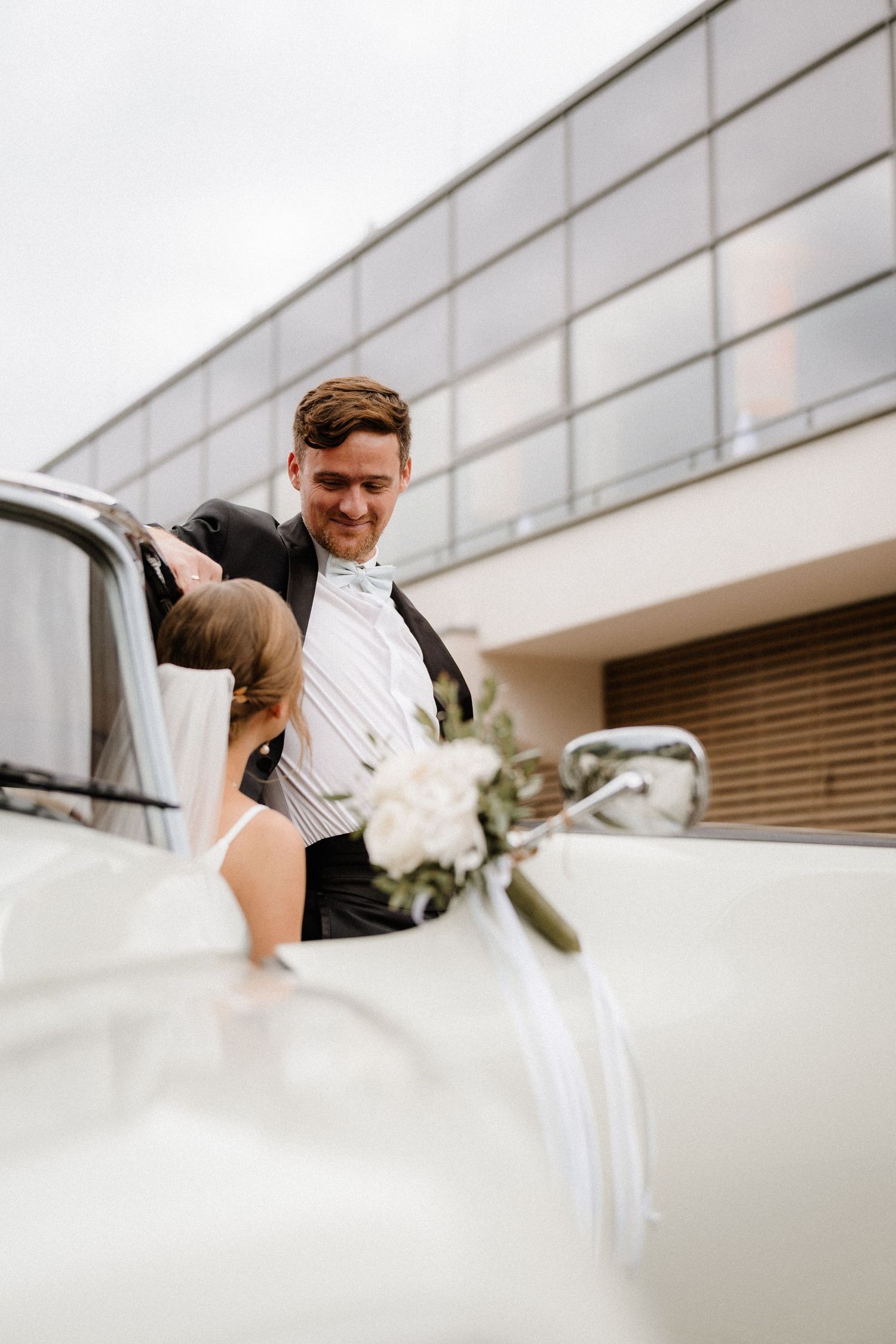 Couple shares romantic moment near modern building exterior with white wedding dress flowing in breeze.