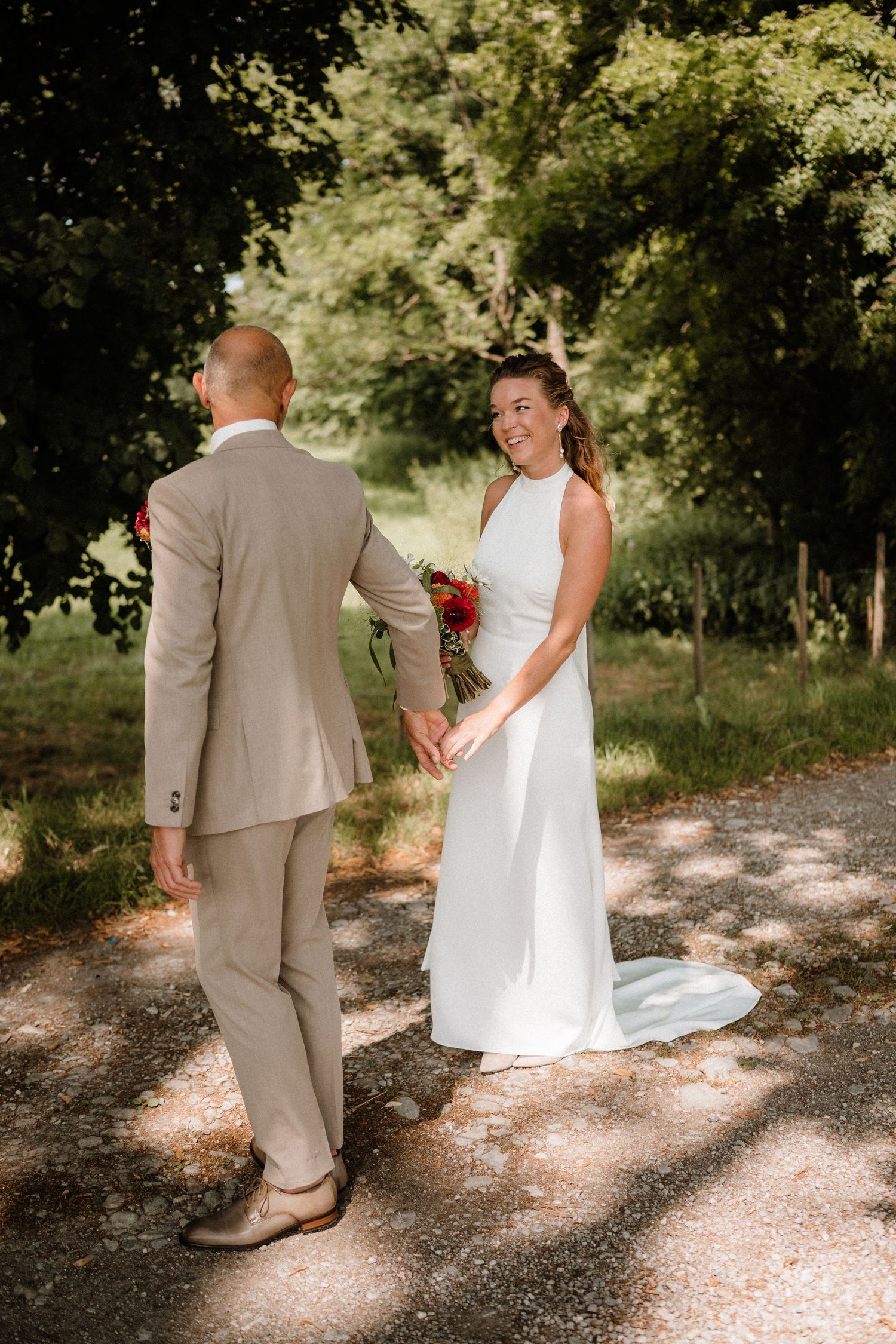 A couple shares a romantic moment on a sunlit gravel path surrounded by trees.