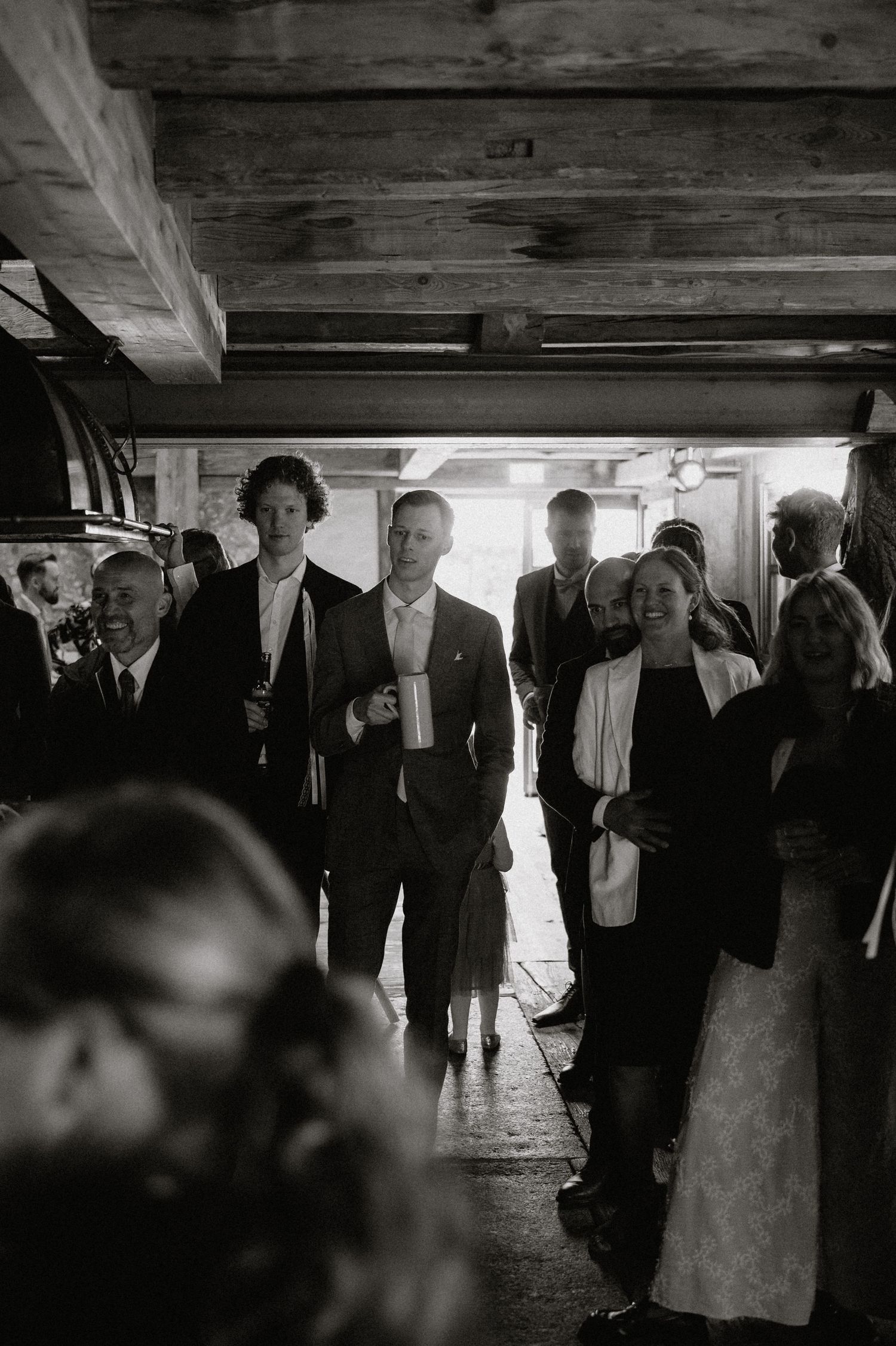 Black and white photo of wedding guests mingling in a rustic wooden interior.