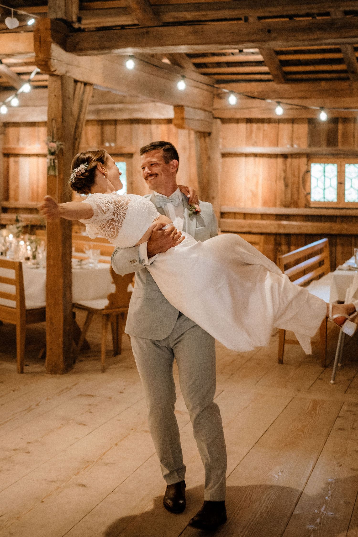 Dancing couple spins across rustic wooden floor under warm lighting in barn venue.