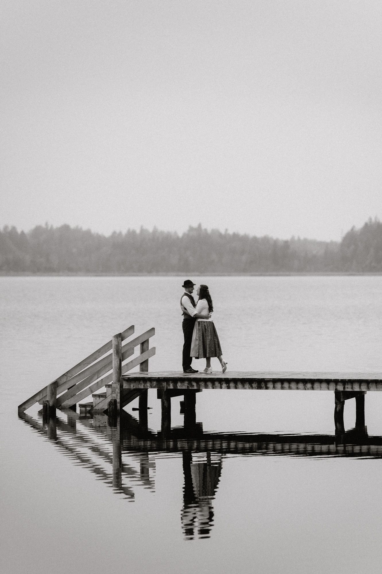 Ein romantisches Paar umarmt sich auf einem Holzdock über einem nebligen See in Schwarz-Weiß-Fotografie. Von Dama Fotografie Bad Tölz