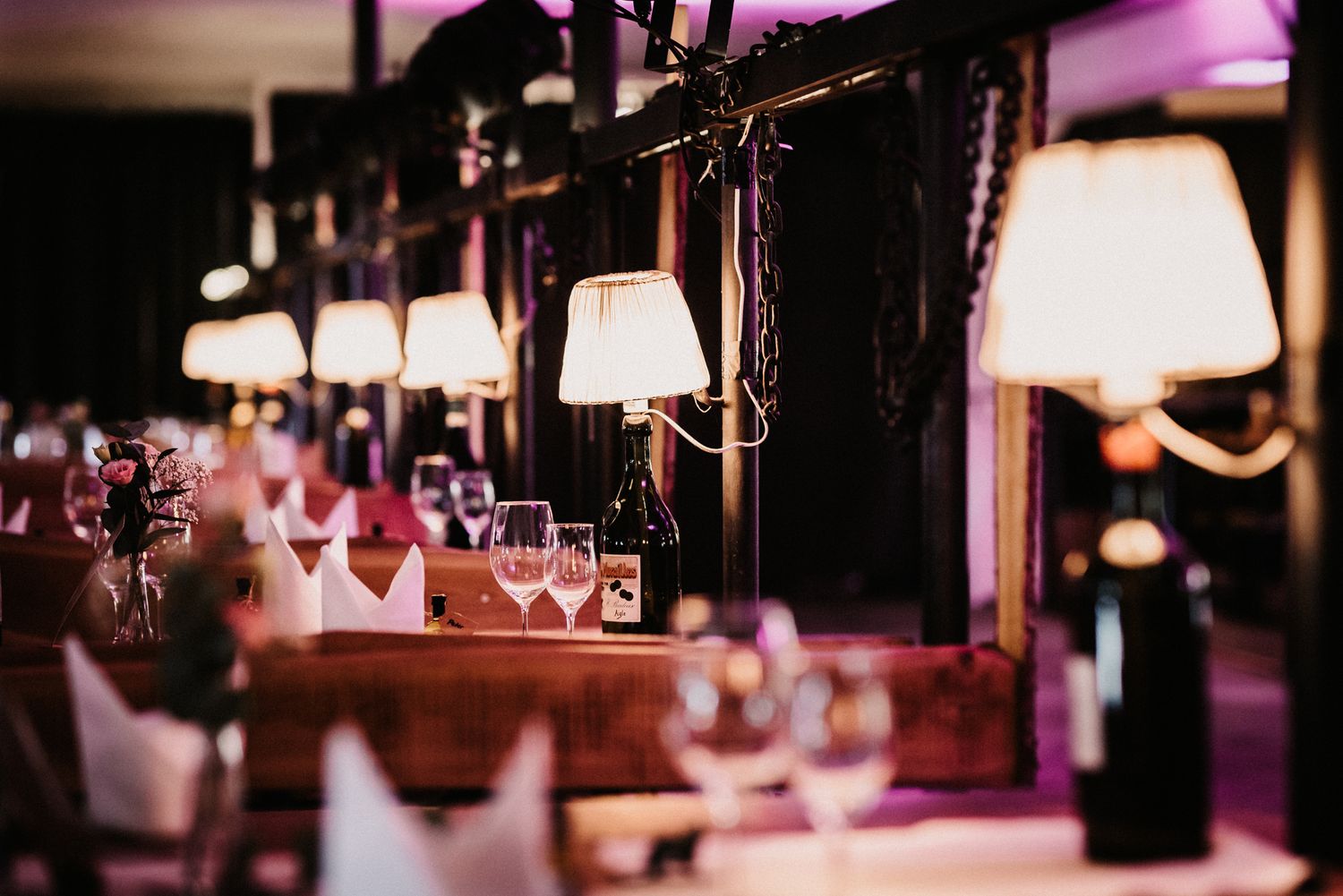 A dimly lit bar interior with purple ambient lighting showcases empty wine glasses and bottles on display.