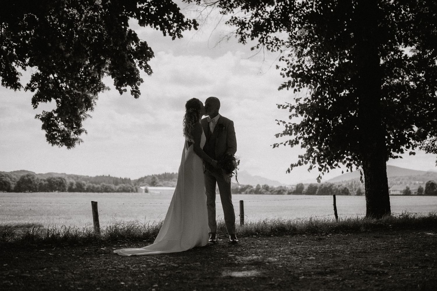 Silhouetten eines Hochzeitspaares stehen unter Bäumen vor einer malerischen Landschaft. Von Dama Fotografie Bad Tölz