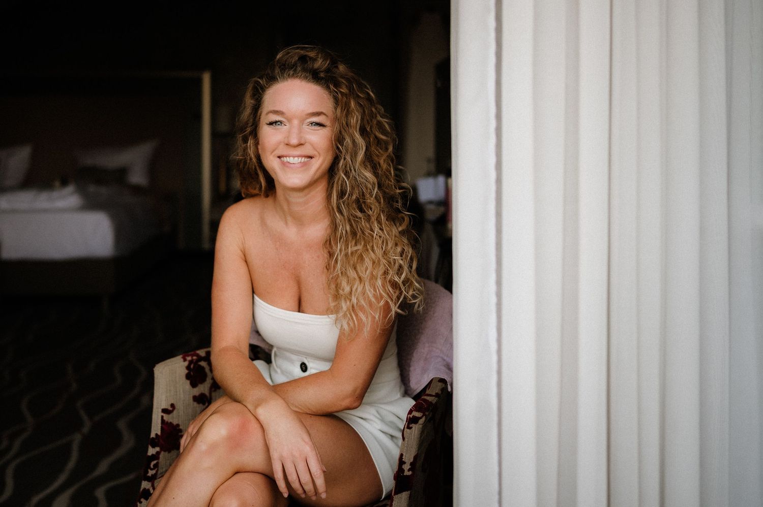 Woman in white dress sitting casually in bedroom setting.