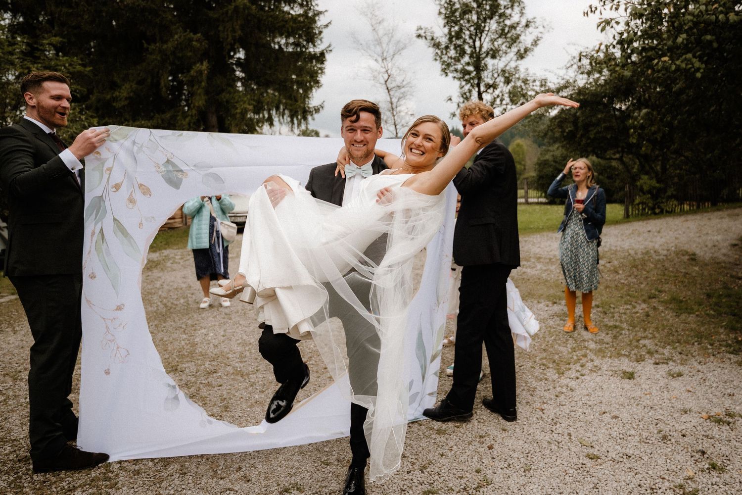 A joyful wedding couple leaps through a white sheet held up by others on a gravel path.