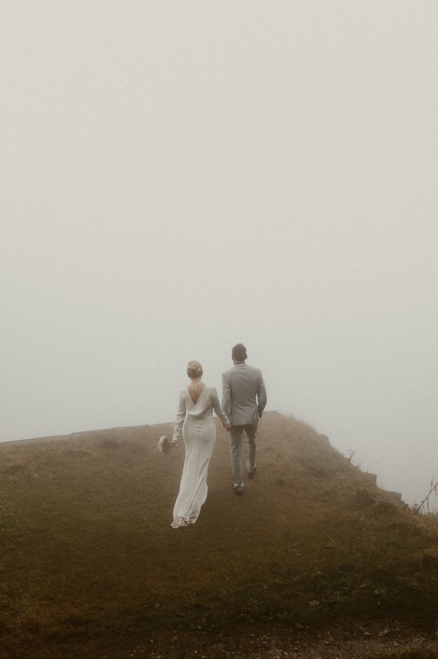 Couple in wedding attire walking along foggy mountain ridge.