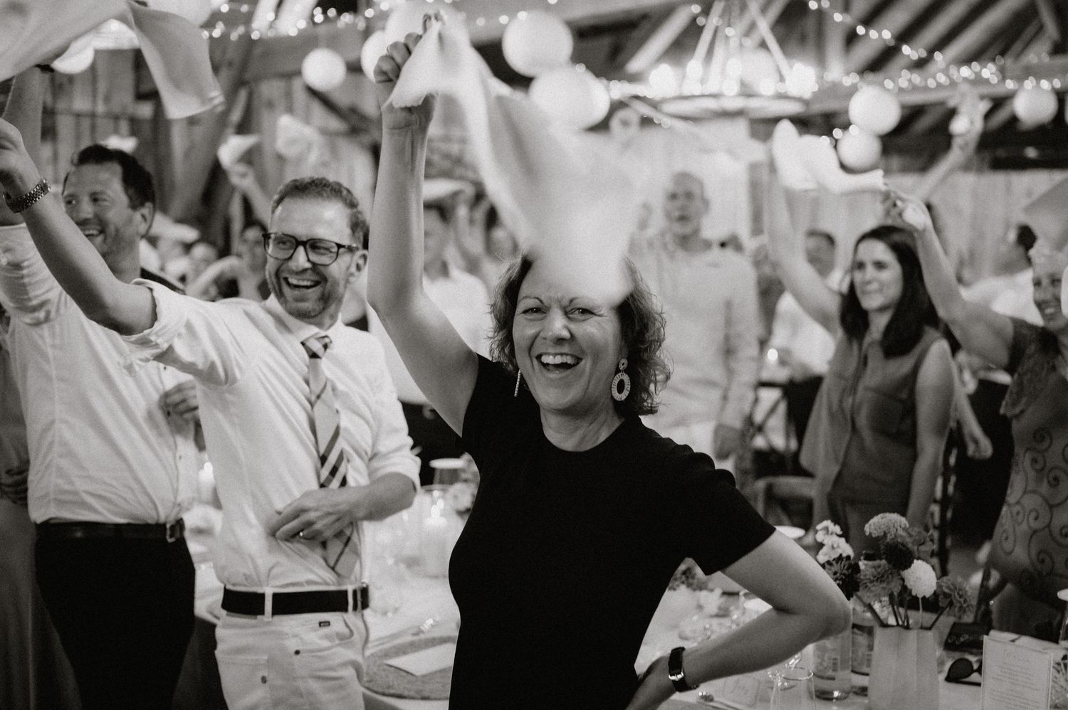 Black and white photo of wedding guests celebrating with arms raised in a barn reception venue.