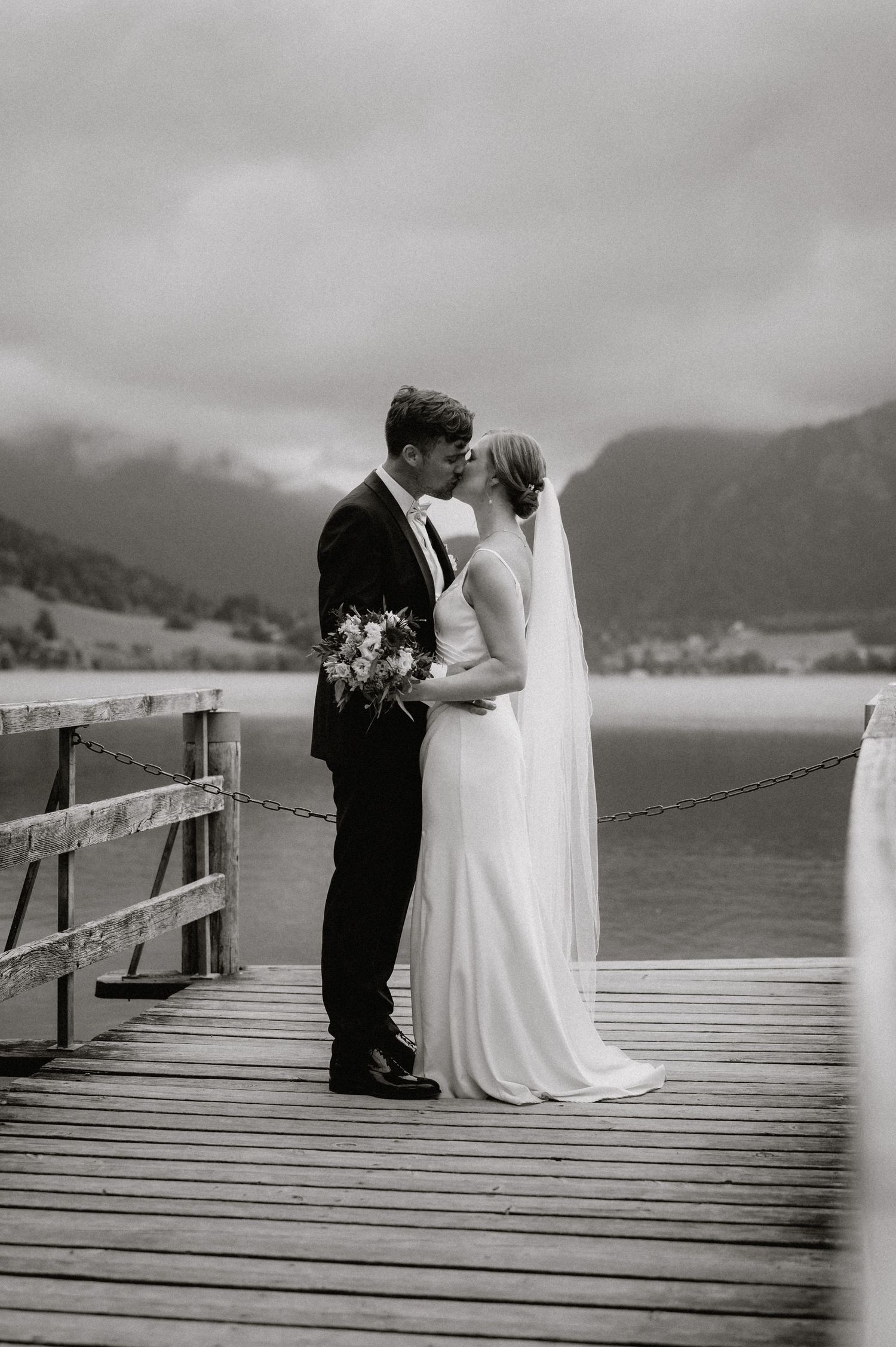 Black and white wedding portrait on wooden pier with mountain lake scenery.