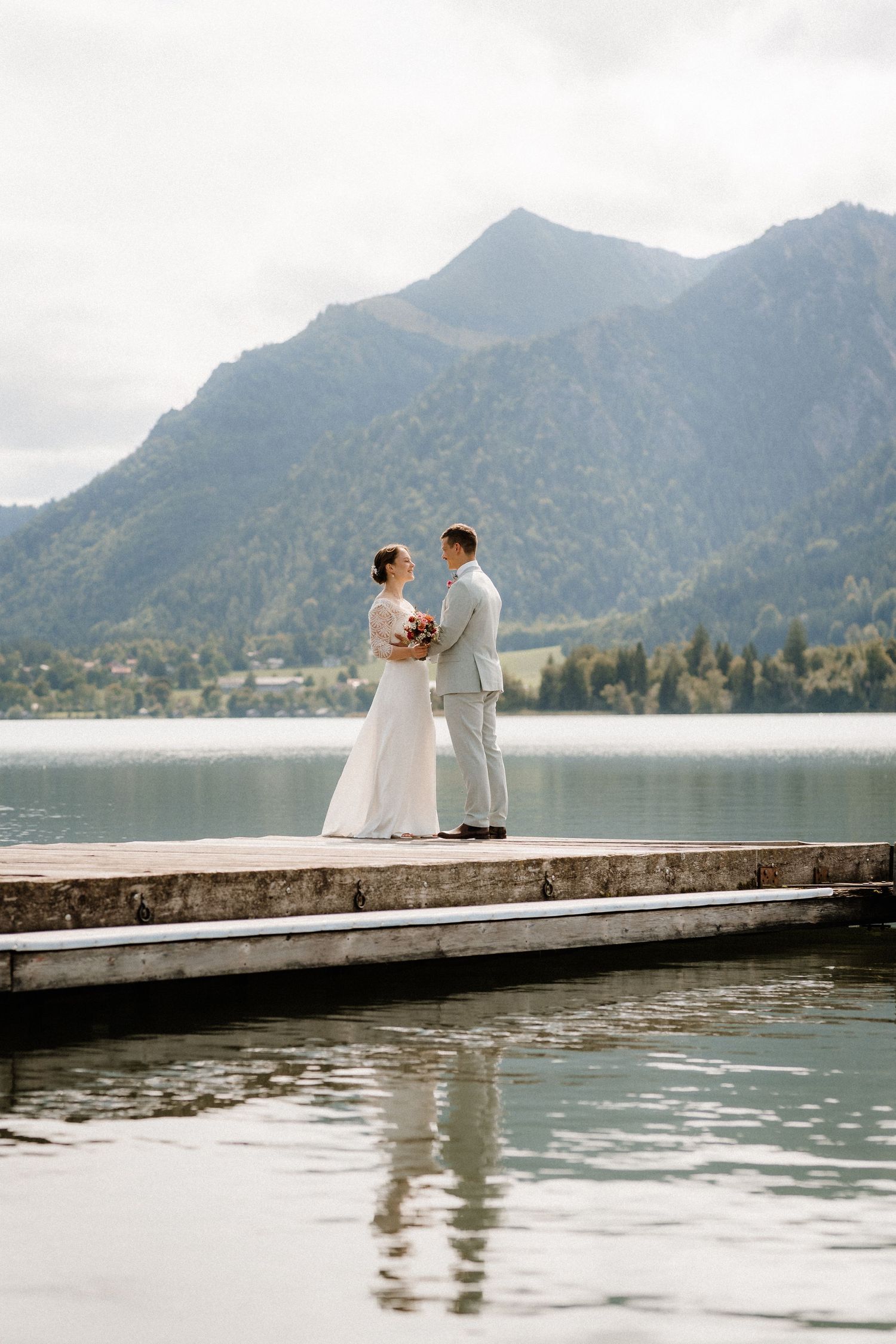 A wedding couple stands on a dock overlooking a mountain lake landscape.