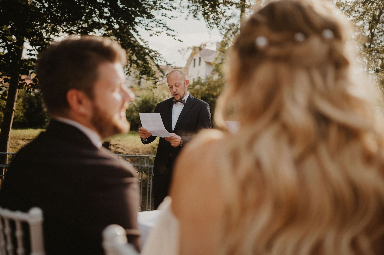 An outdoor wedding ceremony with white chairs and natural lighting filtering through trees.