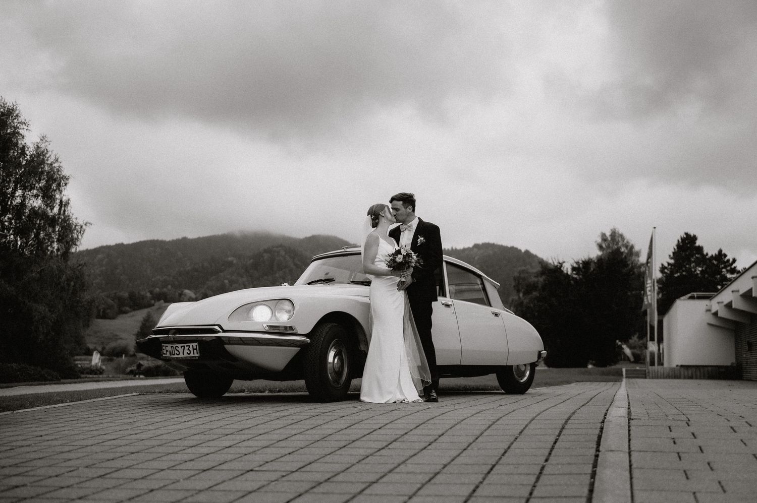 Black and white panoramic shot of wedding couple with vintage car against mountain backdrop.