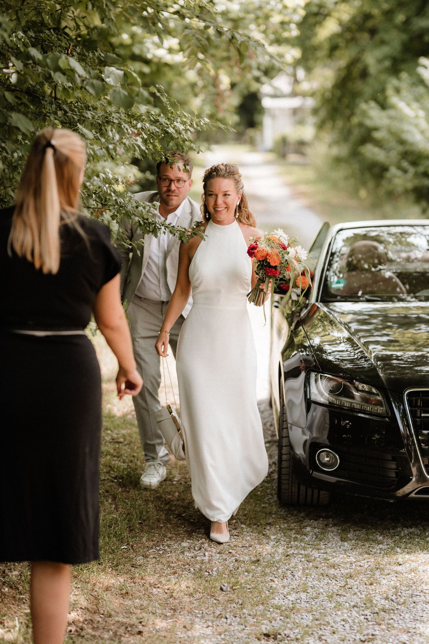 Wedding party gathered around a black car on a tree-lined gravel path.