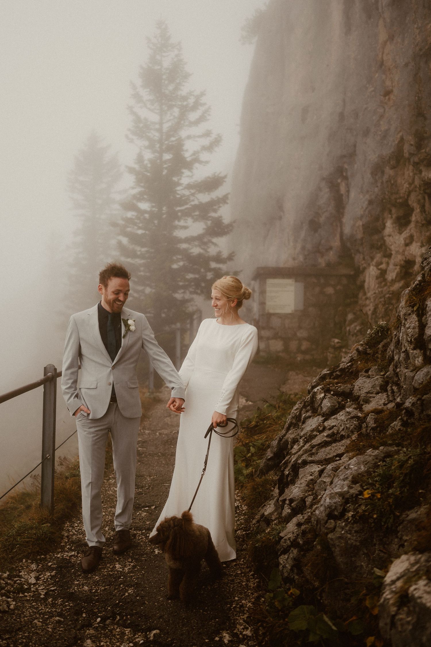Two people in formal attire walk together along a foggy mountain path with rocky cliffs.