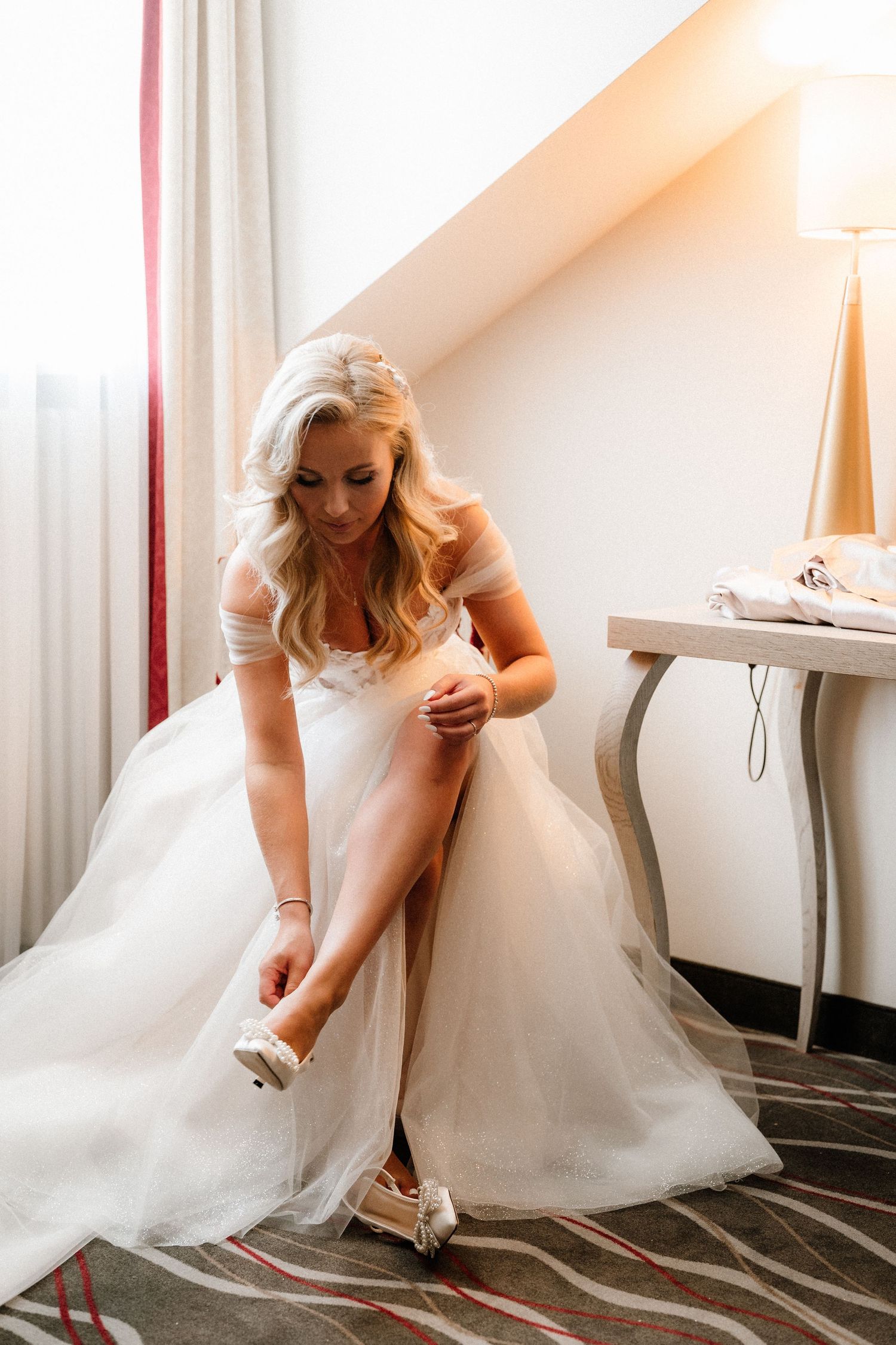 A bride in a white ball gown adjusts her shoe while getting ready in a hotel room.